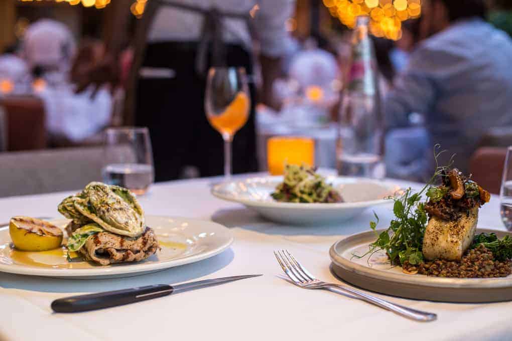 A close-up of a restaurant table with three plated dishes, glasses of water and wine, a bottle, and a lit candle. The background shows people dining and warm string lights out of focus.