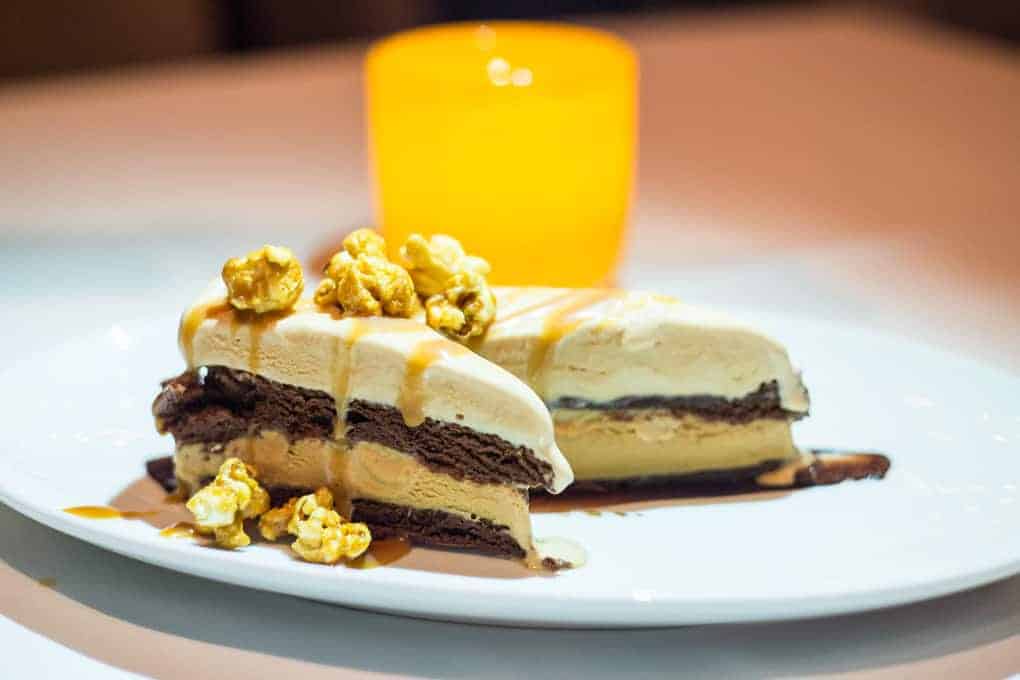 A slice of layered ice cream cake with chocolate and vanilla, topped with caramel drizzle and caramel popcorn, is served on a white plate with an orange cup in the background.