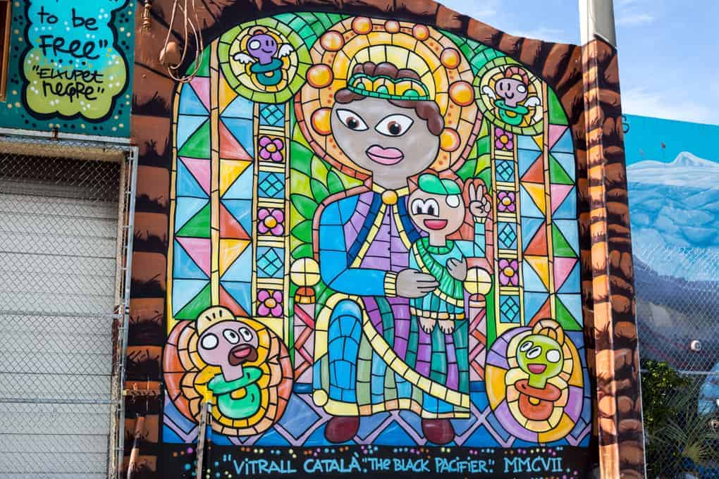 Colorful mural of a crowned figure with four cartoon faces in a stained glass style, framed by geometric patterns.