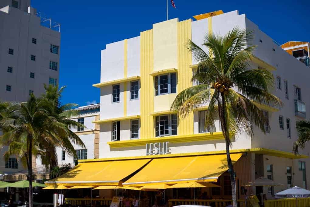 Art Deco hotel with yellow accents and awning, palm trees in front, under a clear blue sky.
