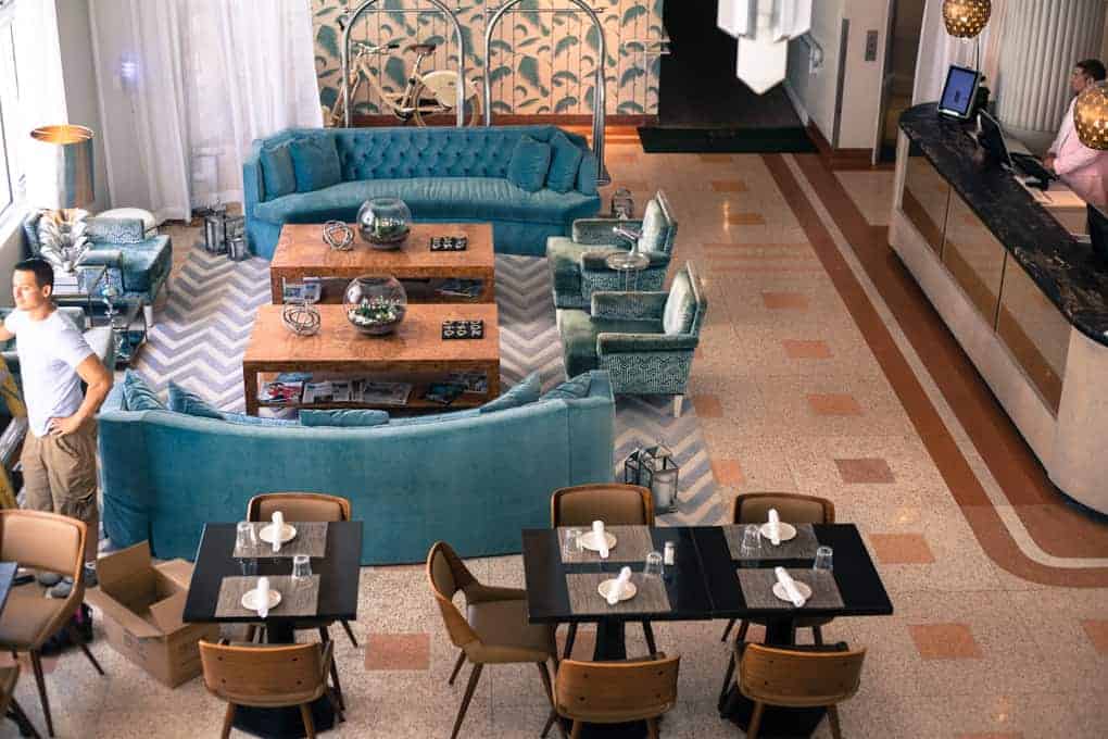 A stylish hotel lobby with teal couches, a patterned rug, dining tables, and a reception desk.