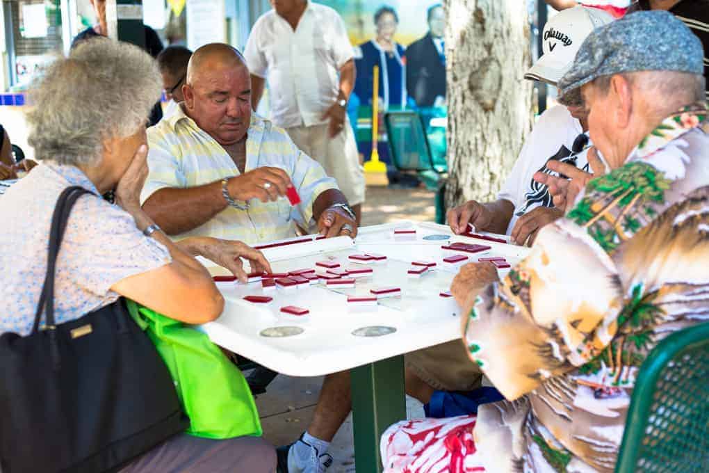 Four elderly people play dominoes at an outdoor table, with others standing in the background.