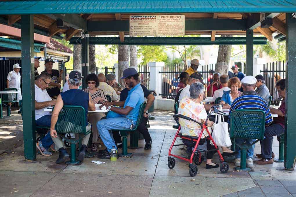 People of various ages sit at tables under a pavilion, playing games and socializing in a park setting.