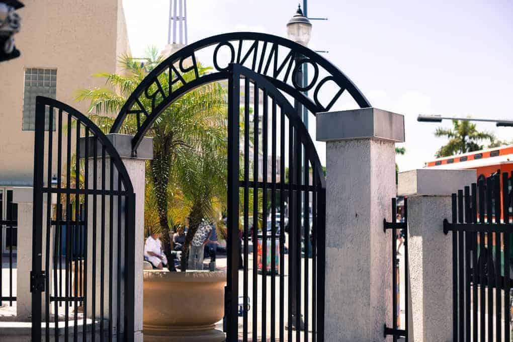 Black iron gates open at the entrance of Domino Park, with a palm tree visible inside.