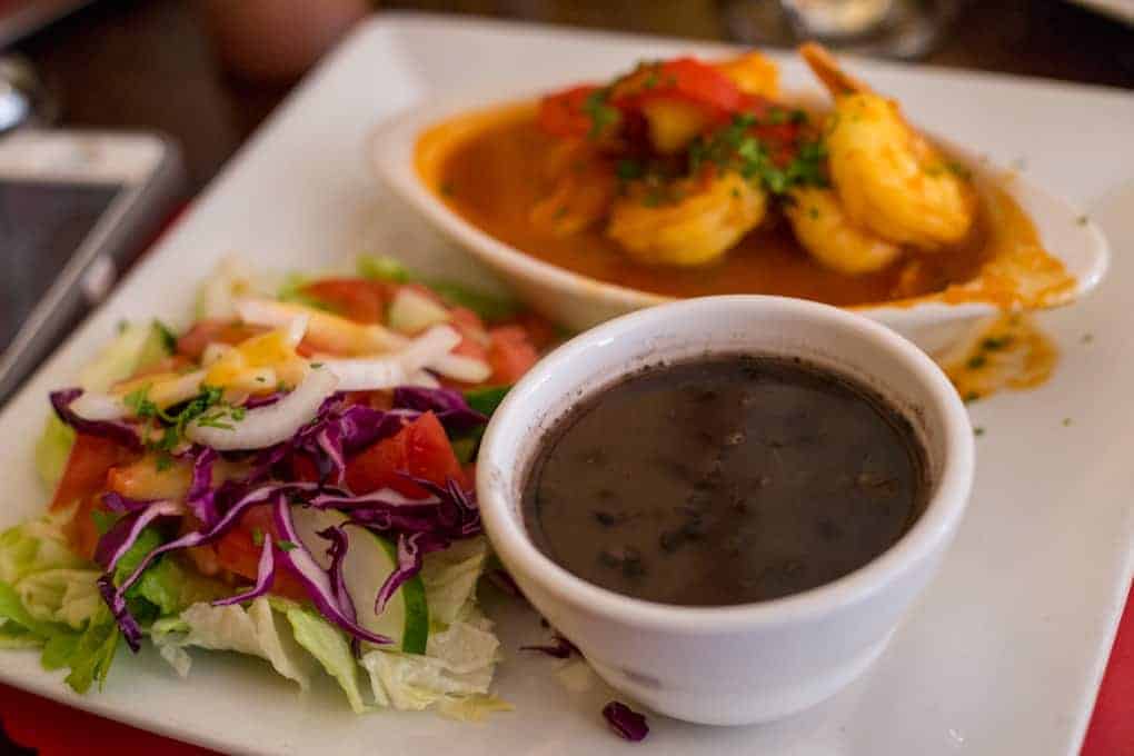 A plate with shrimp in sauce, a cup of black beans, and a fresh salad with lettuce and tomatoes.