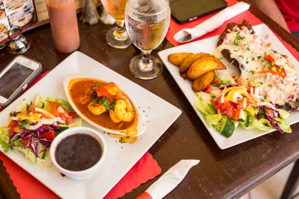 Two plates of colorful food with salad, beans, shrimp, plantains, and drinks on a restaurant table.