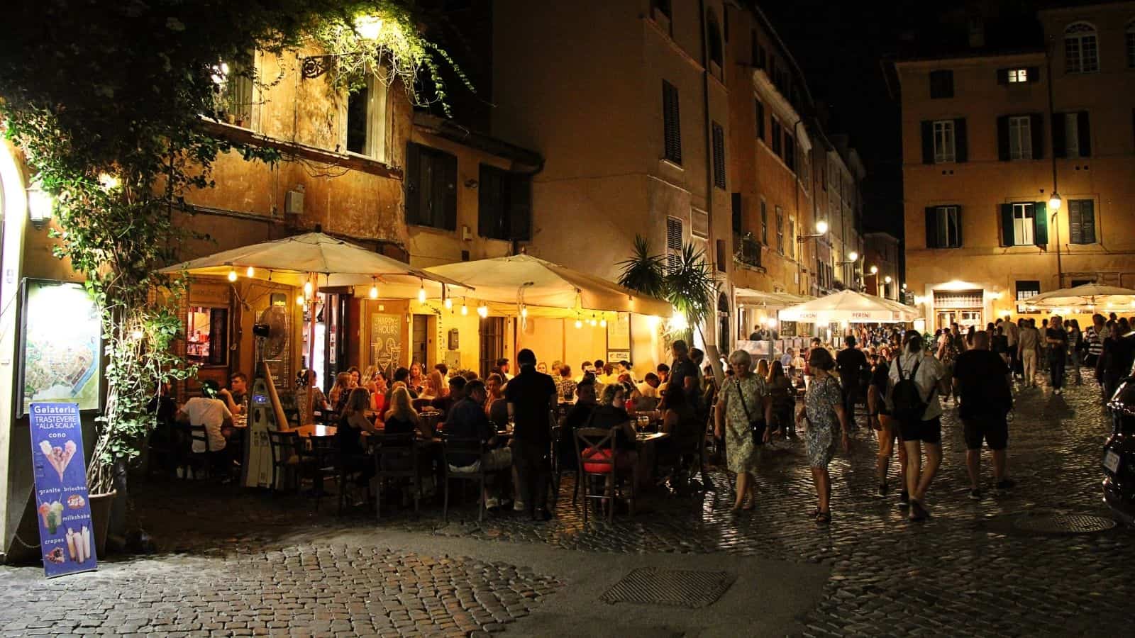 People dine at outdoor tables under umbrellas at a busy restaurant on a cobblestone street at night. Warm lights illuminate the scene, as first-time travelers observe European habits while strolling along the lively, bustling street.
