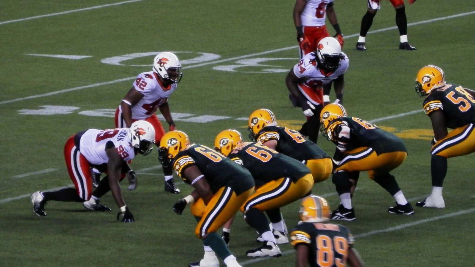 Football players line up on the field before a play. The team in green and yellow prepares to snap the ball while the opposing team in white and red is on defense. Yard lines and hash marks are visible on the turf.