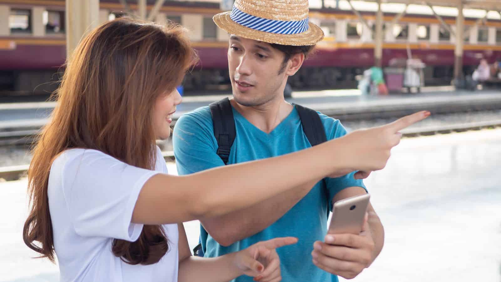 A woman and man at a train station, both pointing and looking in the same direction, holding a phone.