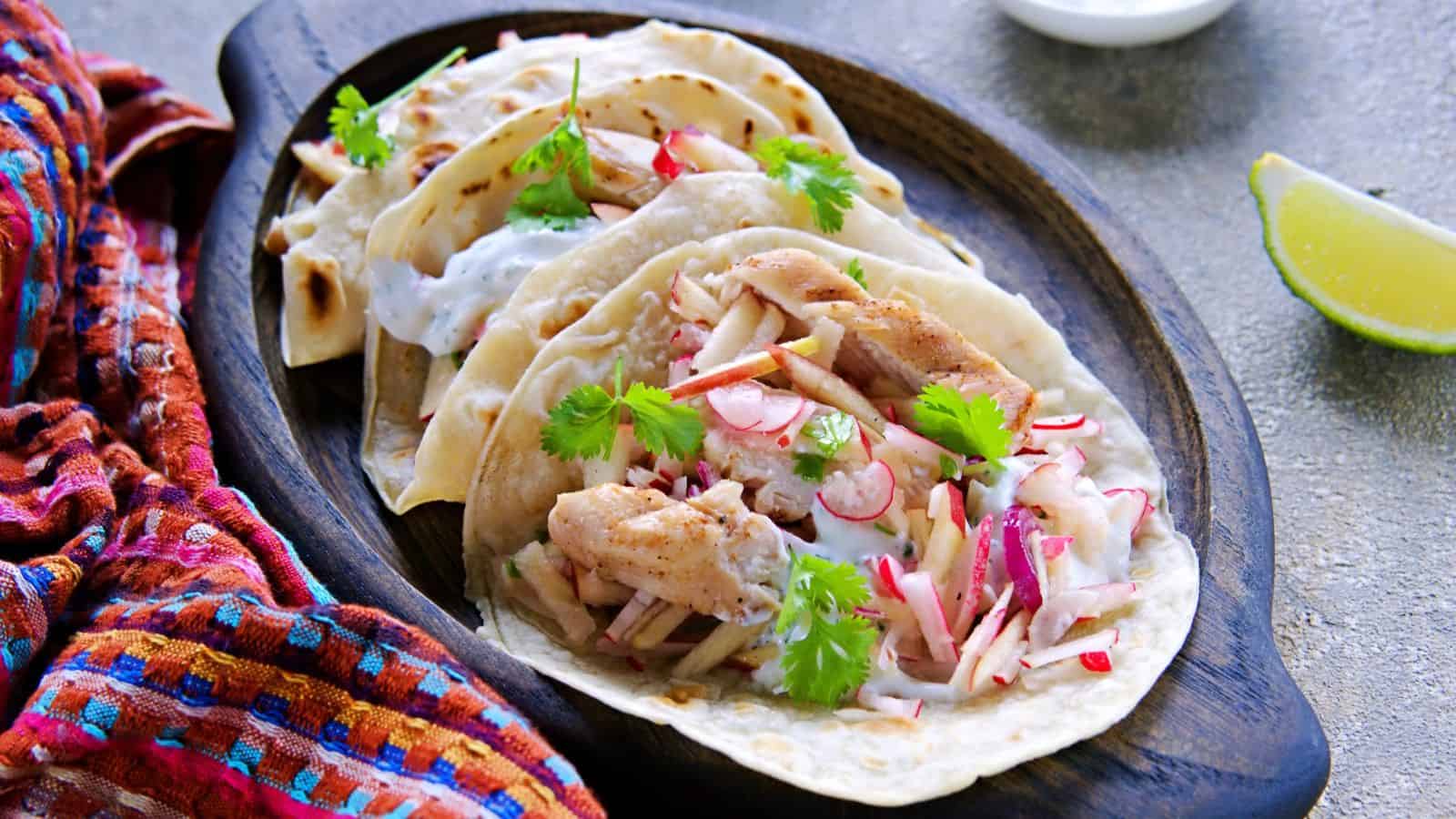 Three grilled fish tacos with shredded vegetables, fresh cilantro, and white sauce are served on a dark wooden platter—making you jealous of U.S. things other countries can only dream of recreating.