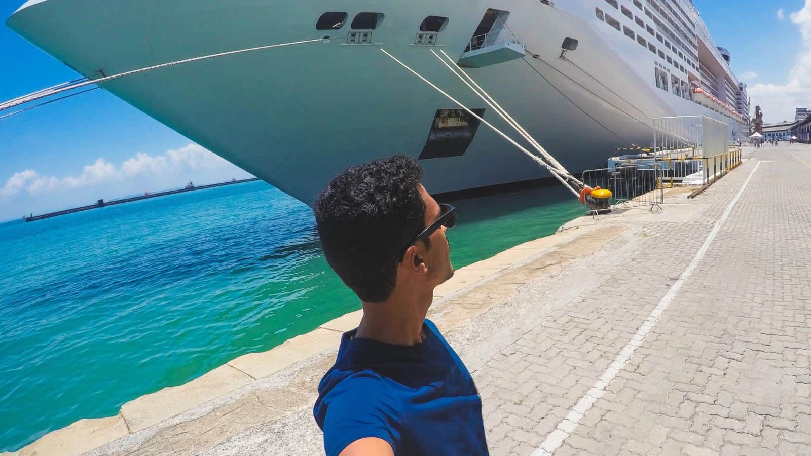 A person wearing sunglasses and a blue shirt takes a selfie near a large white cruise ship docked at a port, with clear turquoise water and a paved walkway in the background.