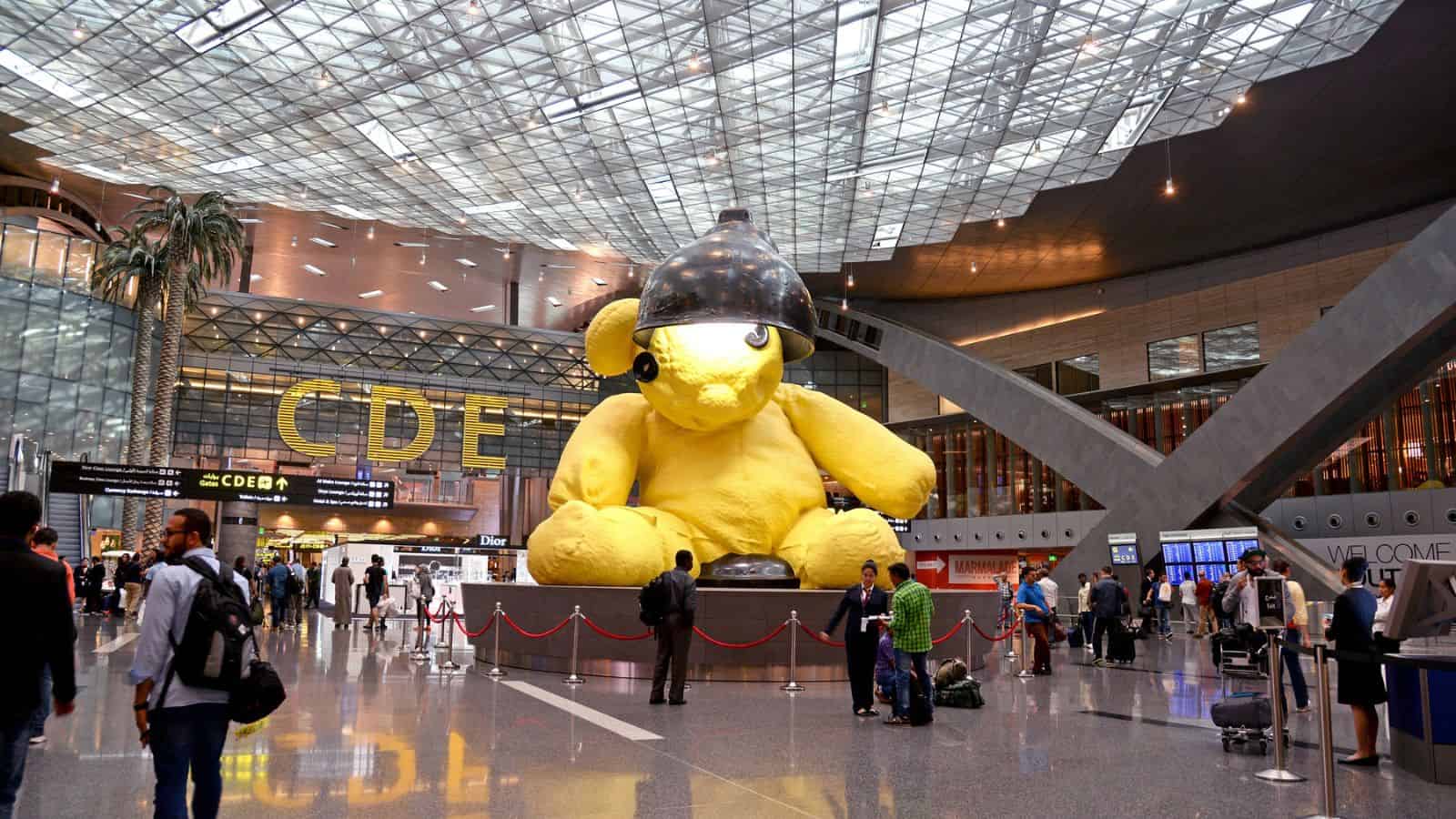 Travelers walk through an airport terminal featuring a large yellow teddy bear sculpture with a black lamp on its head. Signs indicate gates C, D, and E, and the ceiling is glass with a geometric pattern.