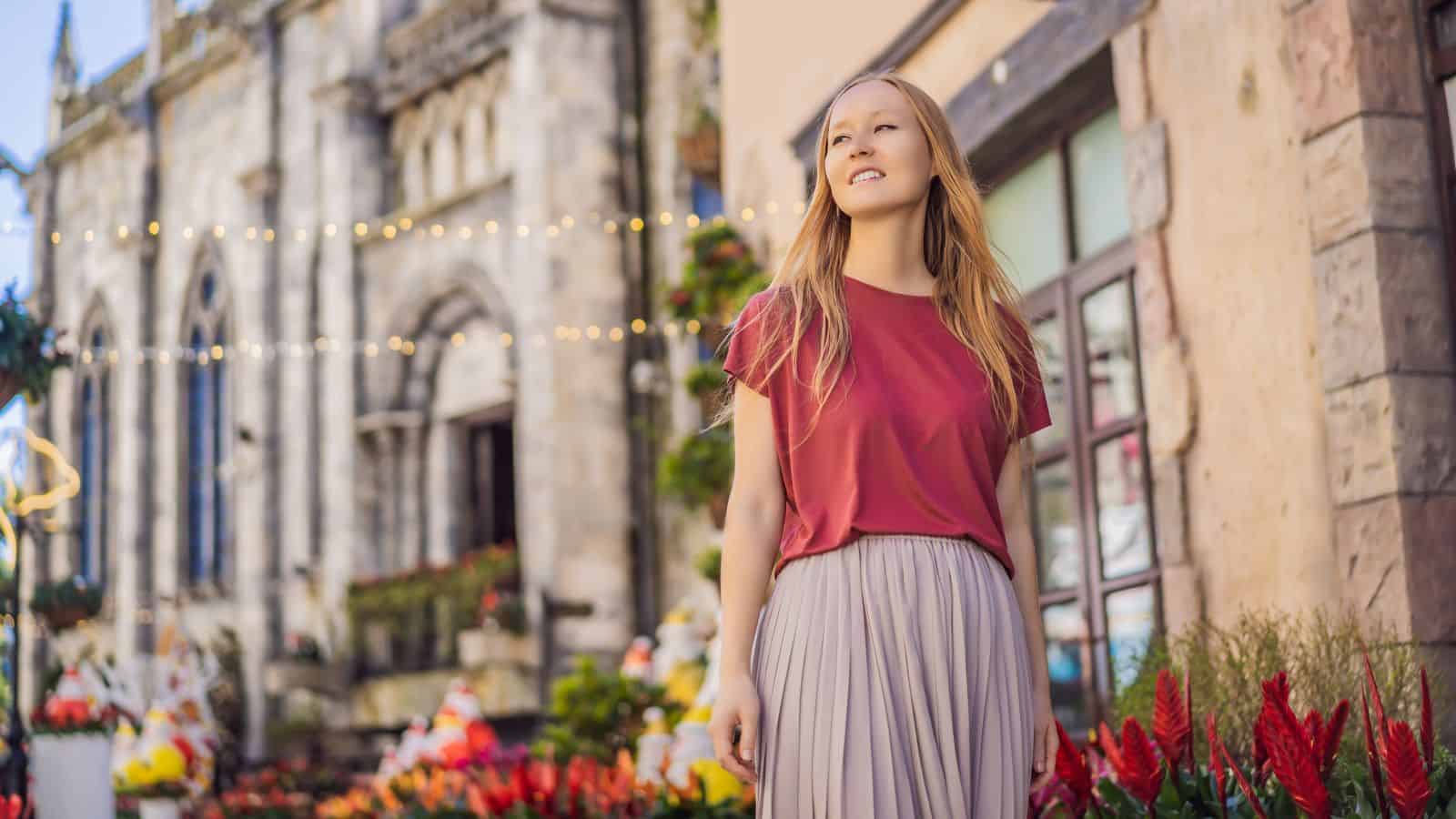 A woman in a red top and pleated skirt smiles while walking near flowers and historic buildings.