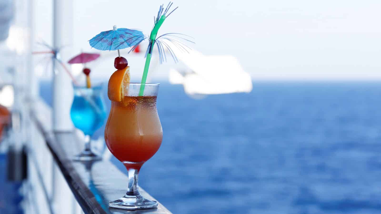 A tropical cocktail with a straw and umbrella on a railing, overlooking the ocean on a sunny day.
