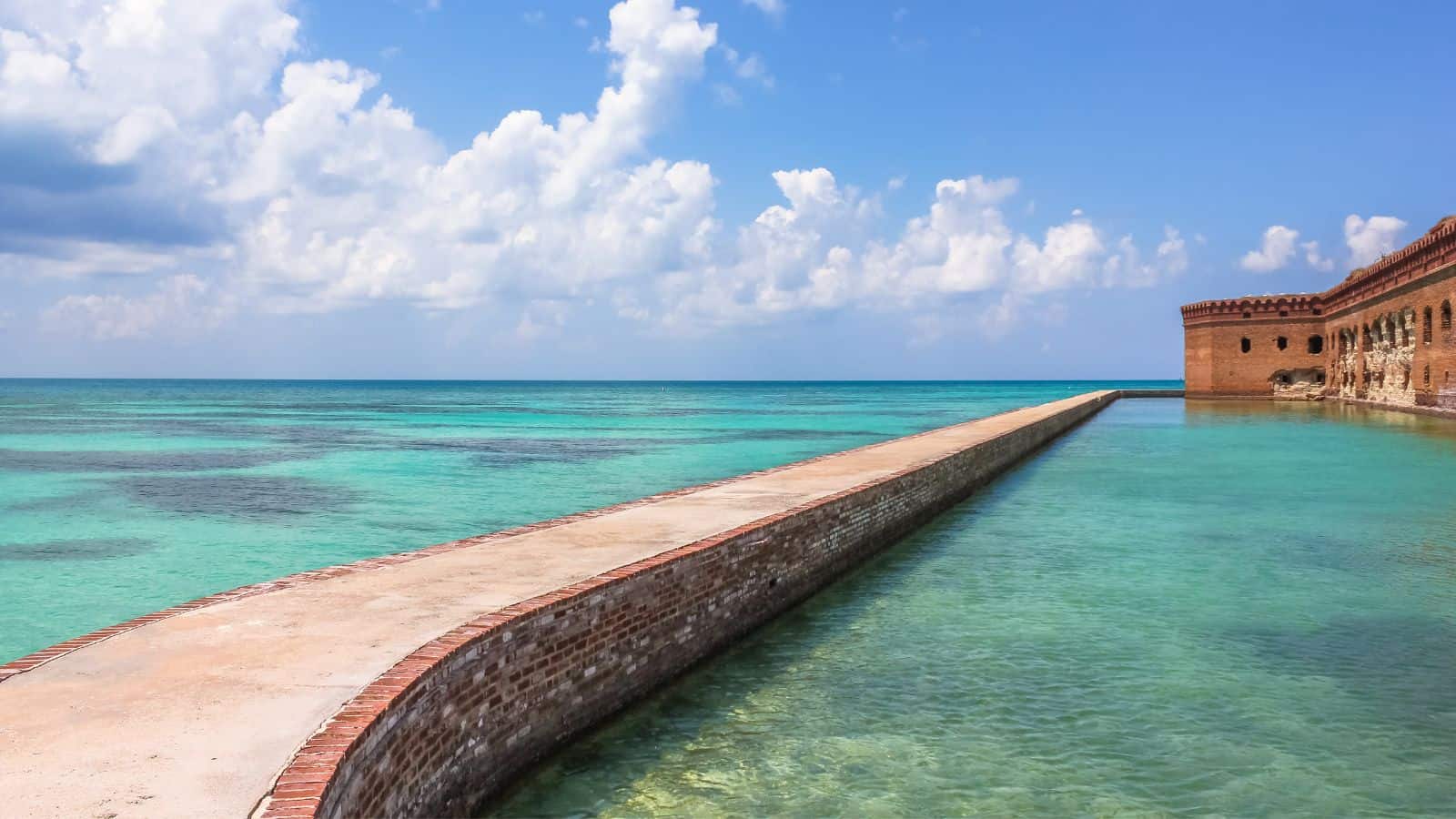A long brick pier extends into clear turquoise waters, with part of a historic brick fort visible on the right and a partly cloudy sky above.