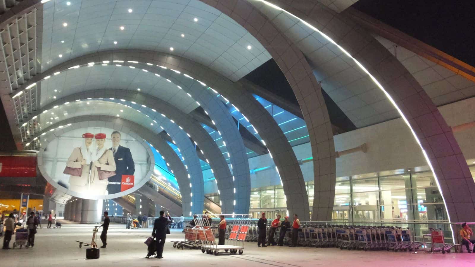 A modern airport terminal at night with large illuminated arches, people walking, and rows of luggage carts. A large advertisement featuring Emirates flight crew is displayed on a circular board.