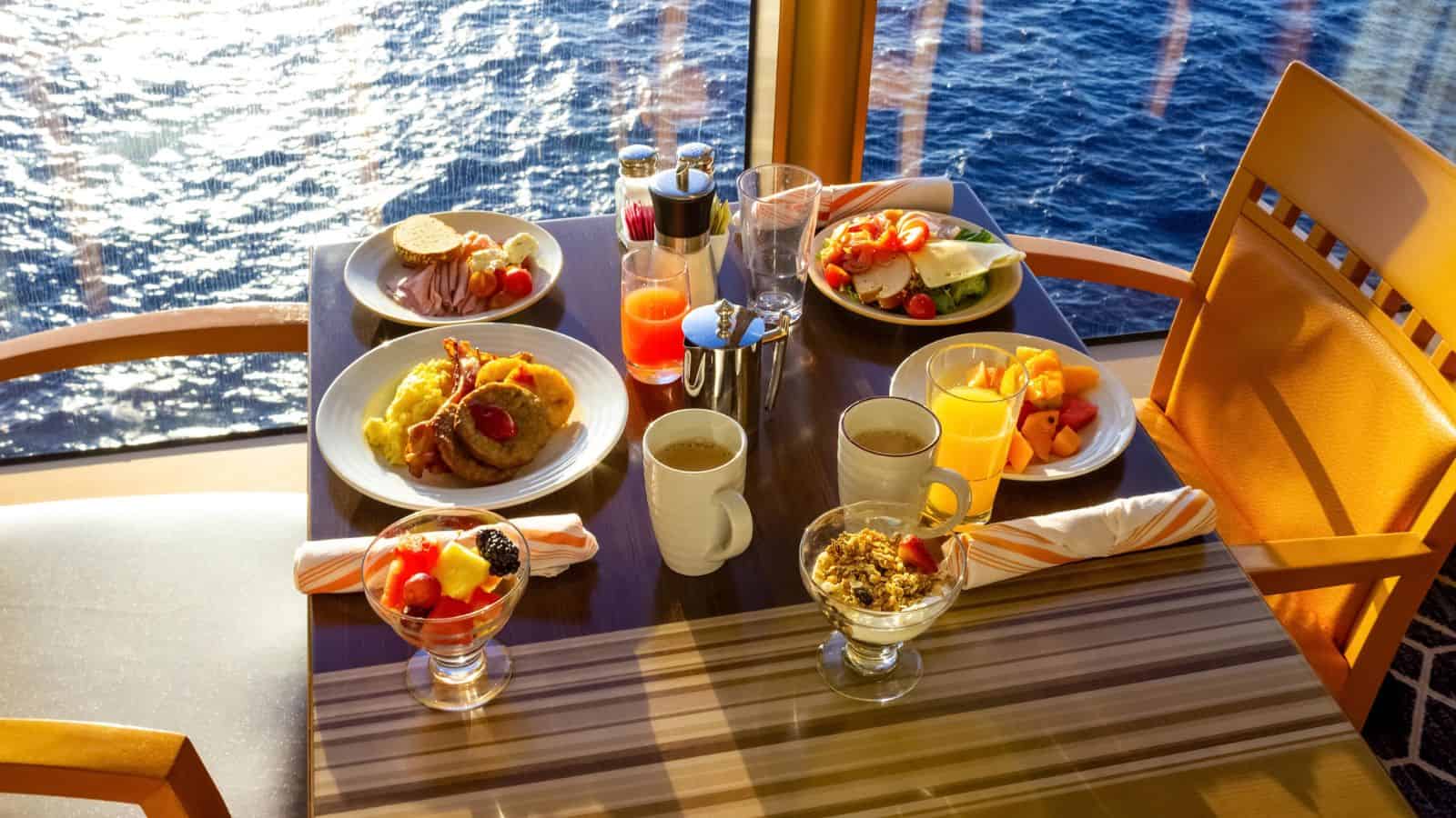 A table set for two with plates of breakfast food, fruit, parfaits, drinks, and coffee, next to a window overlooking the ocean. Sunlight streams onto the table and chairs.