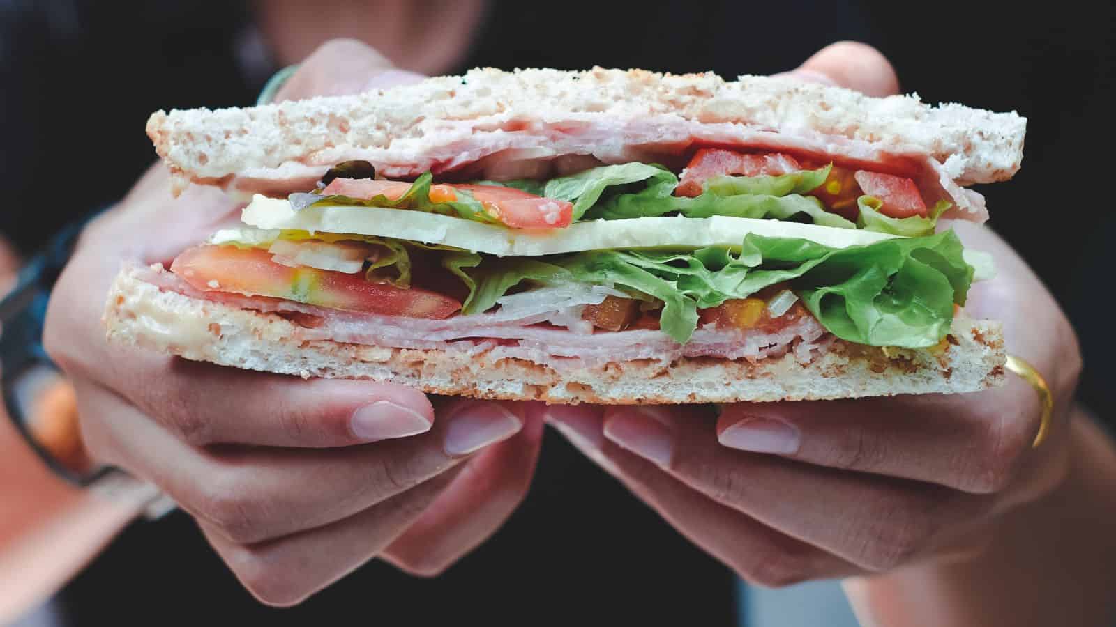 A Paris local holds a sandwich with lettuce, tomato, and sliced meat between two slices of bread—enjoying a simple meal and avoiding common tourist mistakes in Paris.