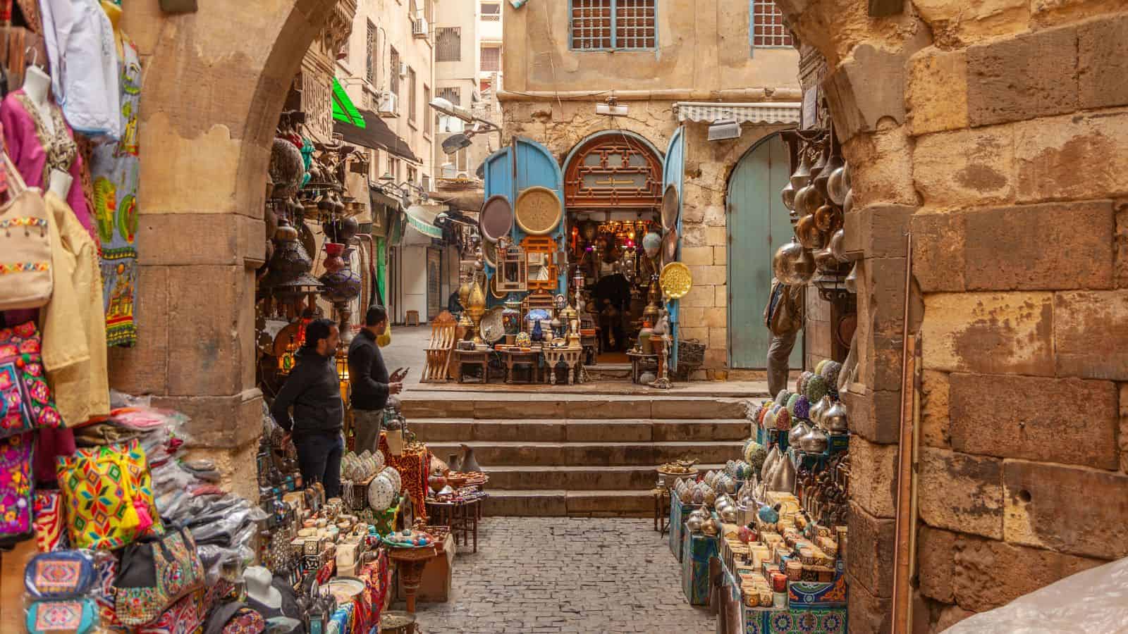 A bustling outdoor market with colorful goods, pottery, and textiles displayed in a narrow stone alleyway.