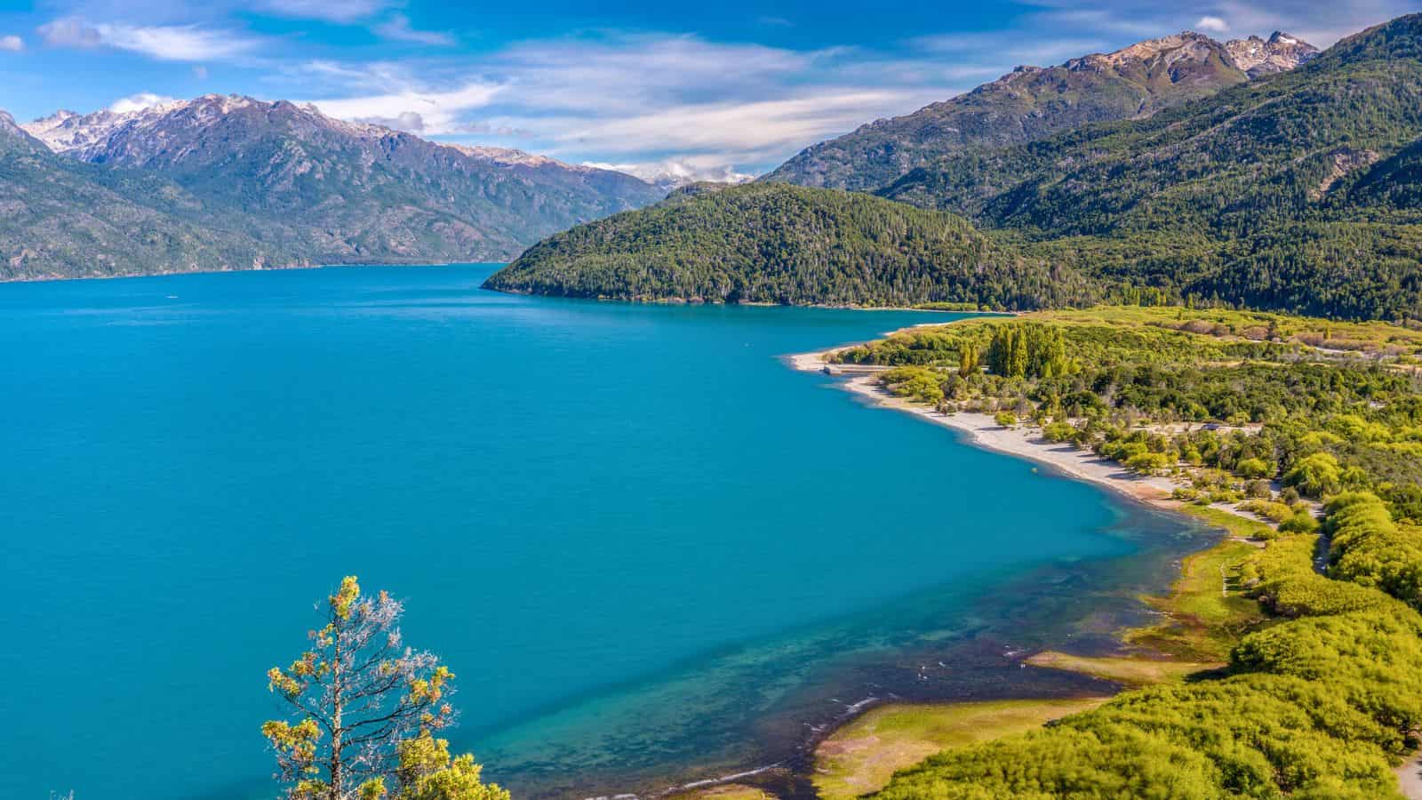 Bright blue lake surrounded by green hills and distant mountains under a partly cloudy sky.