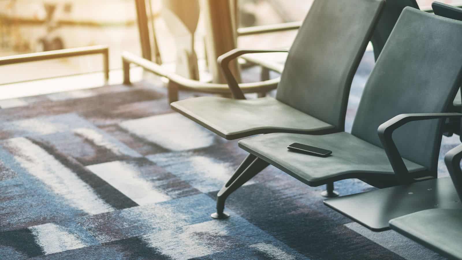 A black smartphone, one of the most commonly lost items in U.S. airports, rests on an empty gray chair. The chairs line up on a blue patterned carpet as sunlight streams through large windows in the background.