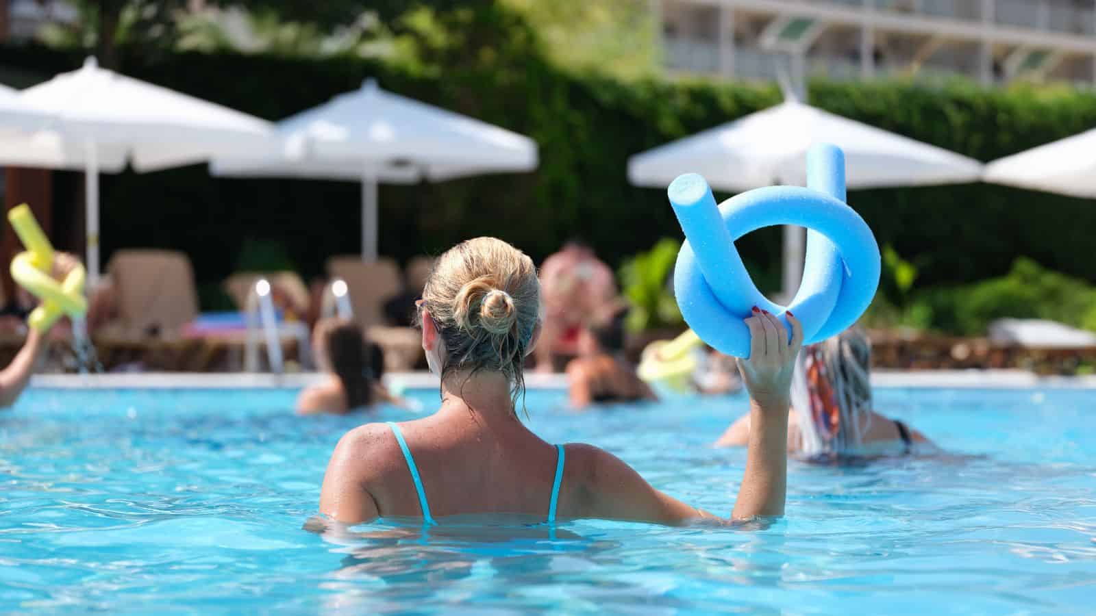 A woman in a blue swimsuit holds a blue pool noodle while standing in a swimming pool with other travelers. White umbrellas and lounge chairs are visible in the background, highlighting one of many vacation reasons to love all-inclusive vacations.