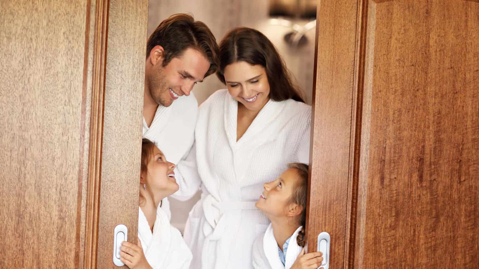 A family of four in white robes smiles at each other while standing between two wooden doors.