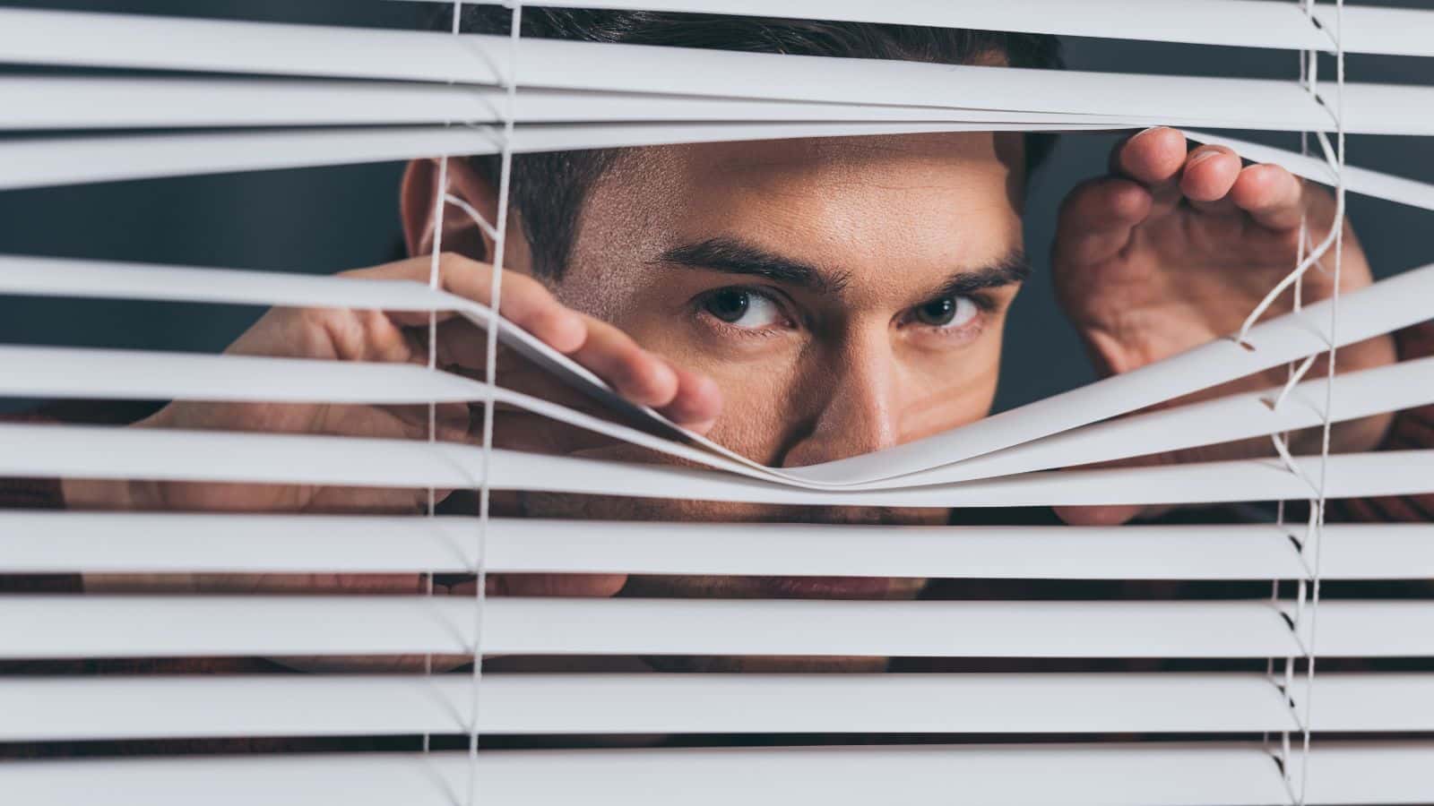 A man peeks through white window blinds with a serious expression on his face.