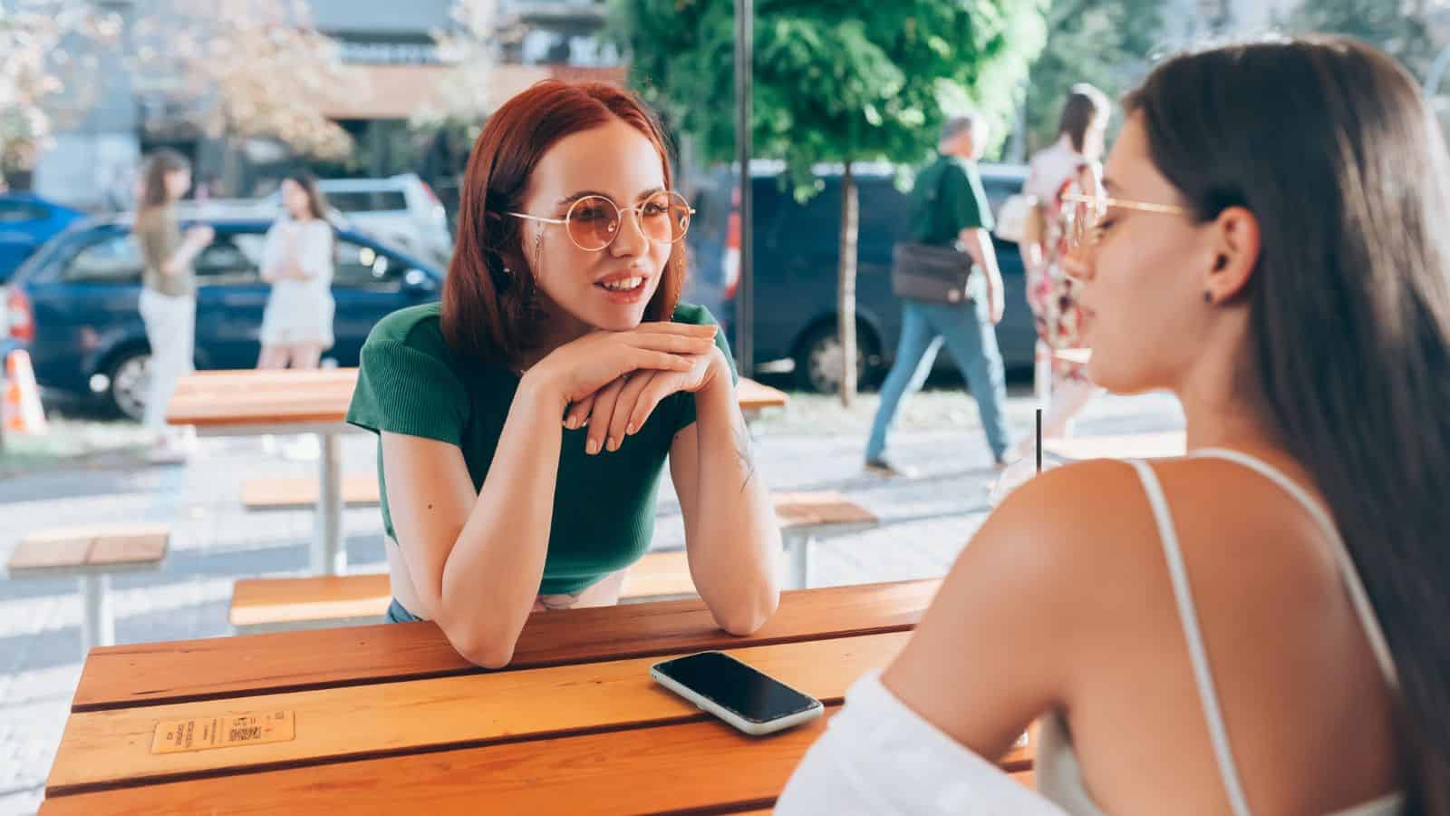 Two women sit at an outdoor table, talking and smiling, with a phone on the table beside them as they share Paris travel tips and insights from Paris locals.