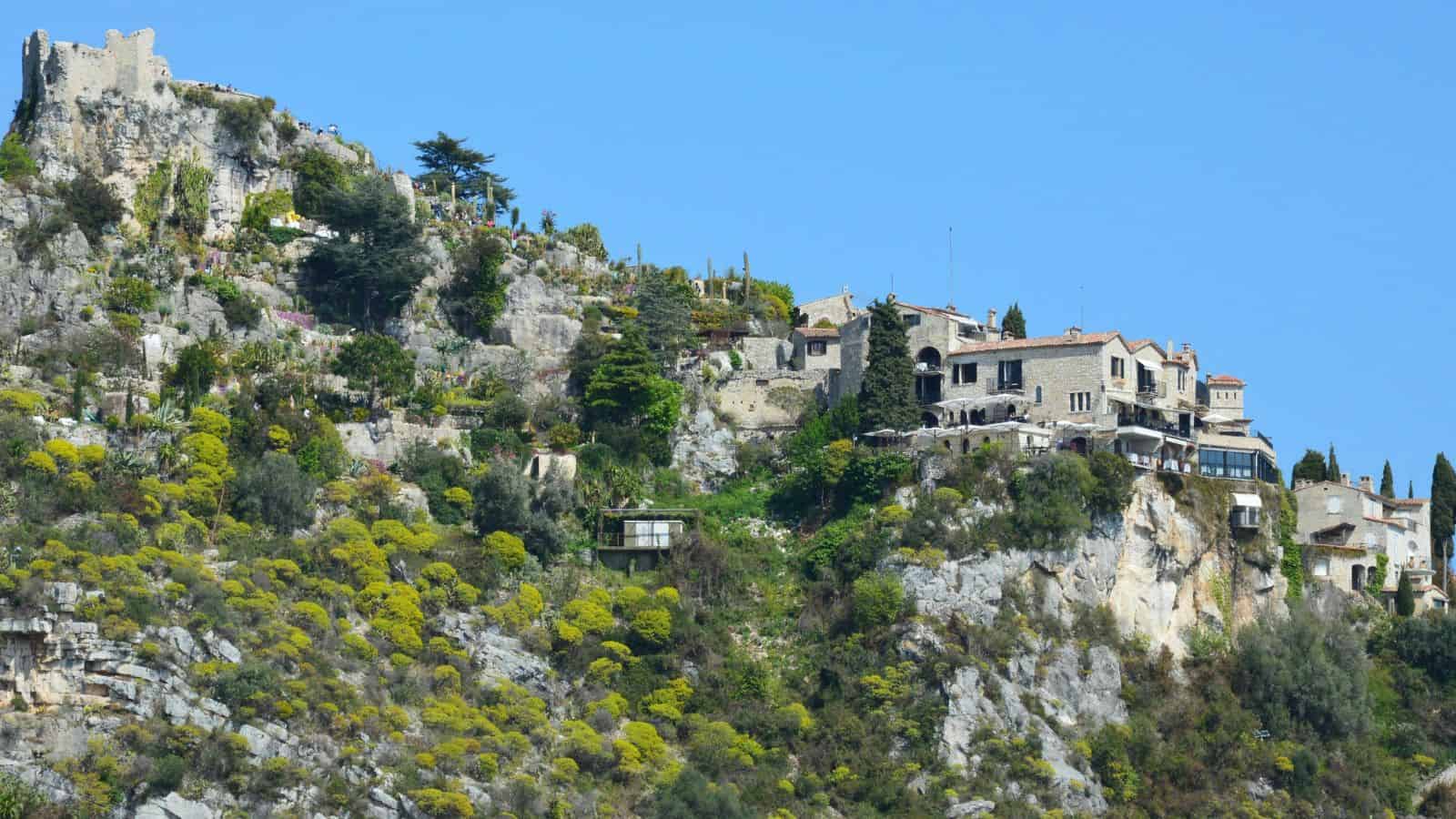 Stone buildings sit atop a rocky, green hillside under a clear blue sky.