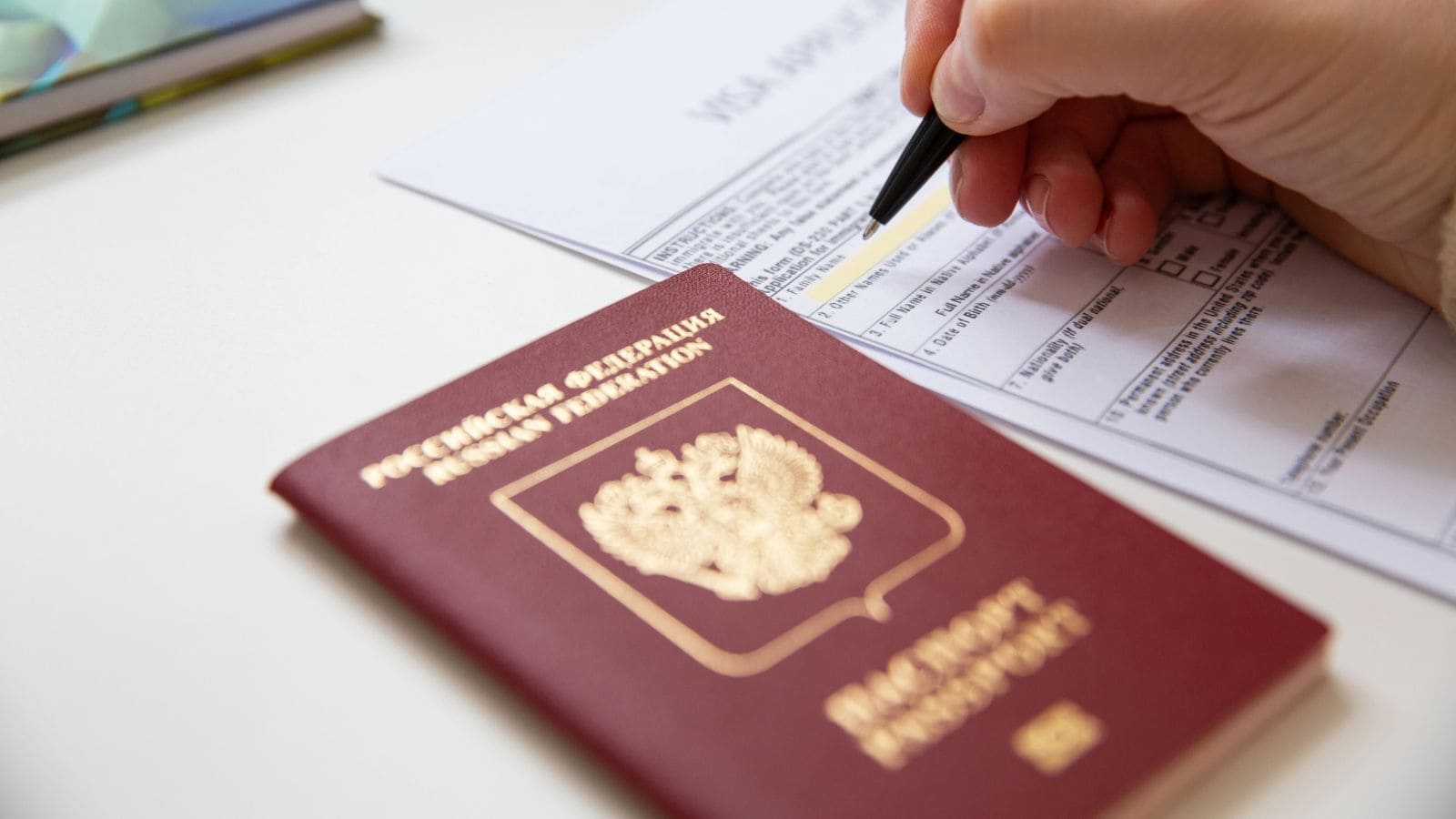 A hand holding a pen fills out a form on a desk. In the foreground, there is a Russian Federation passport with gold text and emblem.