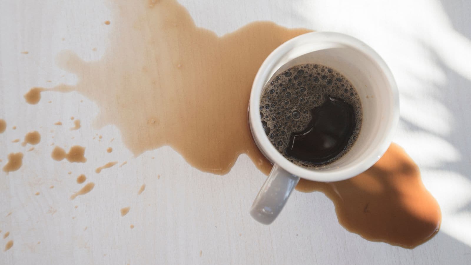 A white mug of coffee tipped over, spilling coffee across a light-colored table.