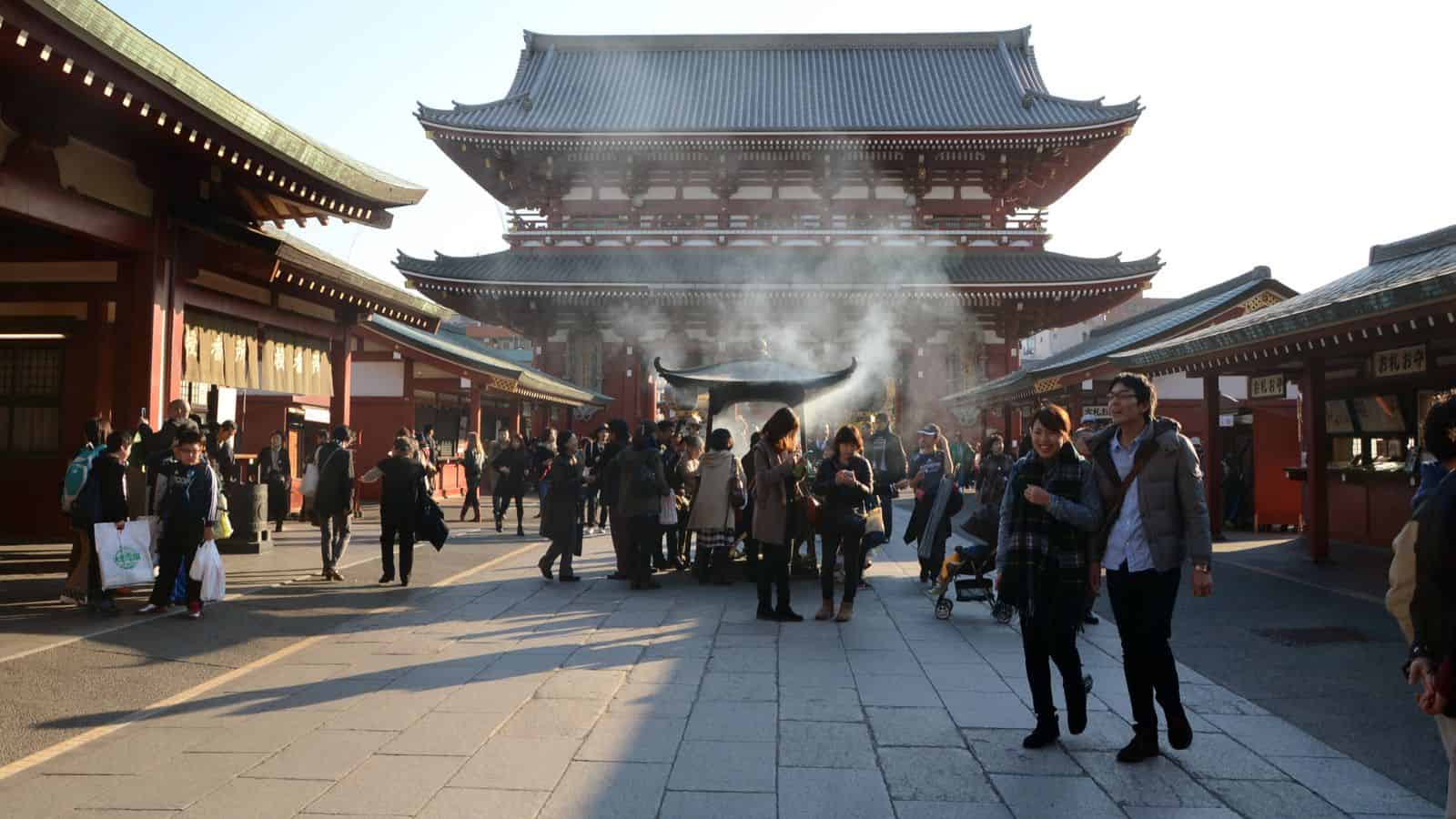 People walking near a smoking incense burner at a traditional Japanese temple on a sunny day.