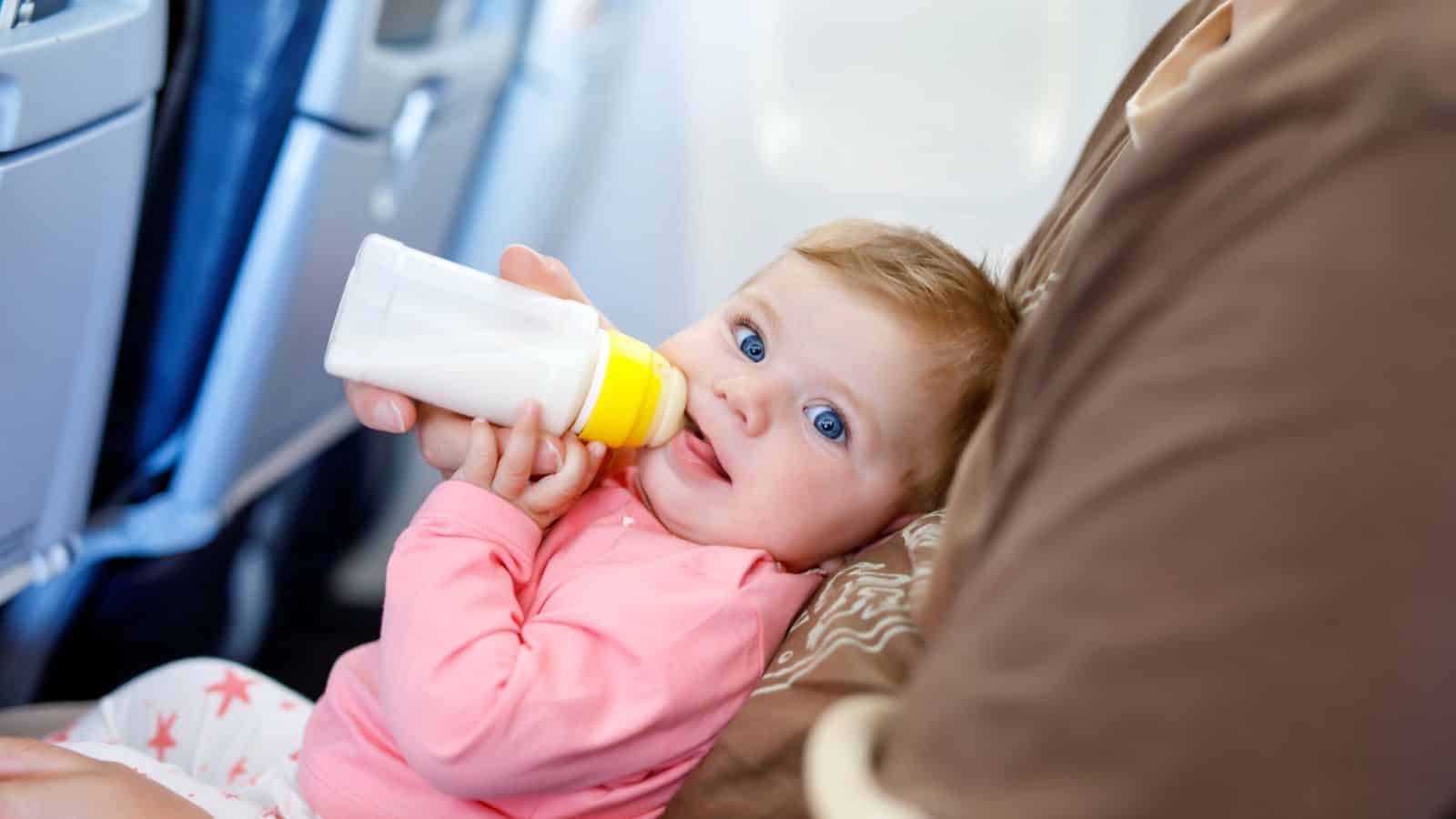 Baby in pink outfit drinking from a bottle while sitting on an adult's lap on an airplane.