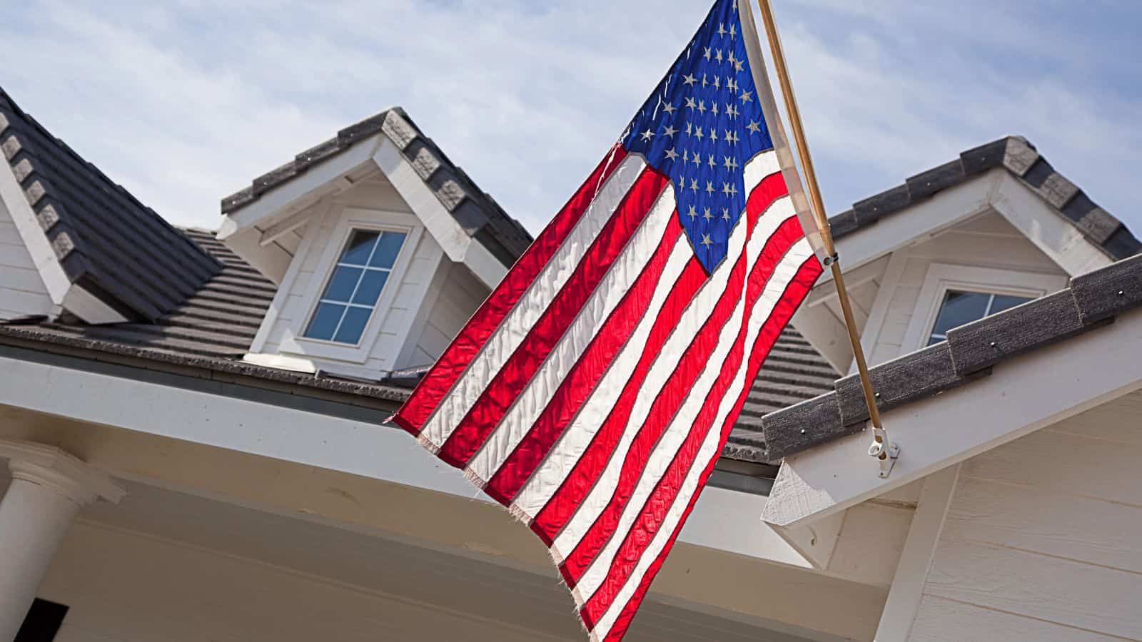 An American flag is mounted on the front of a white house with a tiled roof and dormer windows, under a partly cloudy sky.