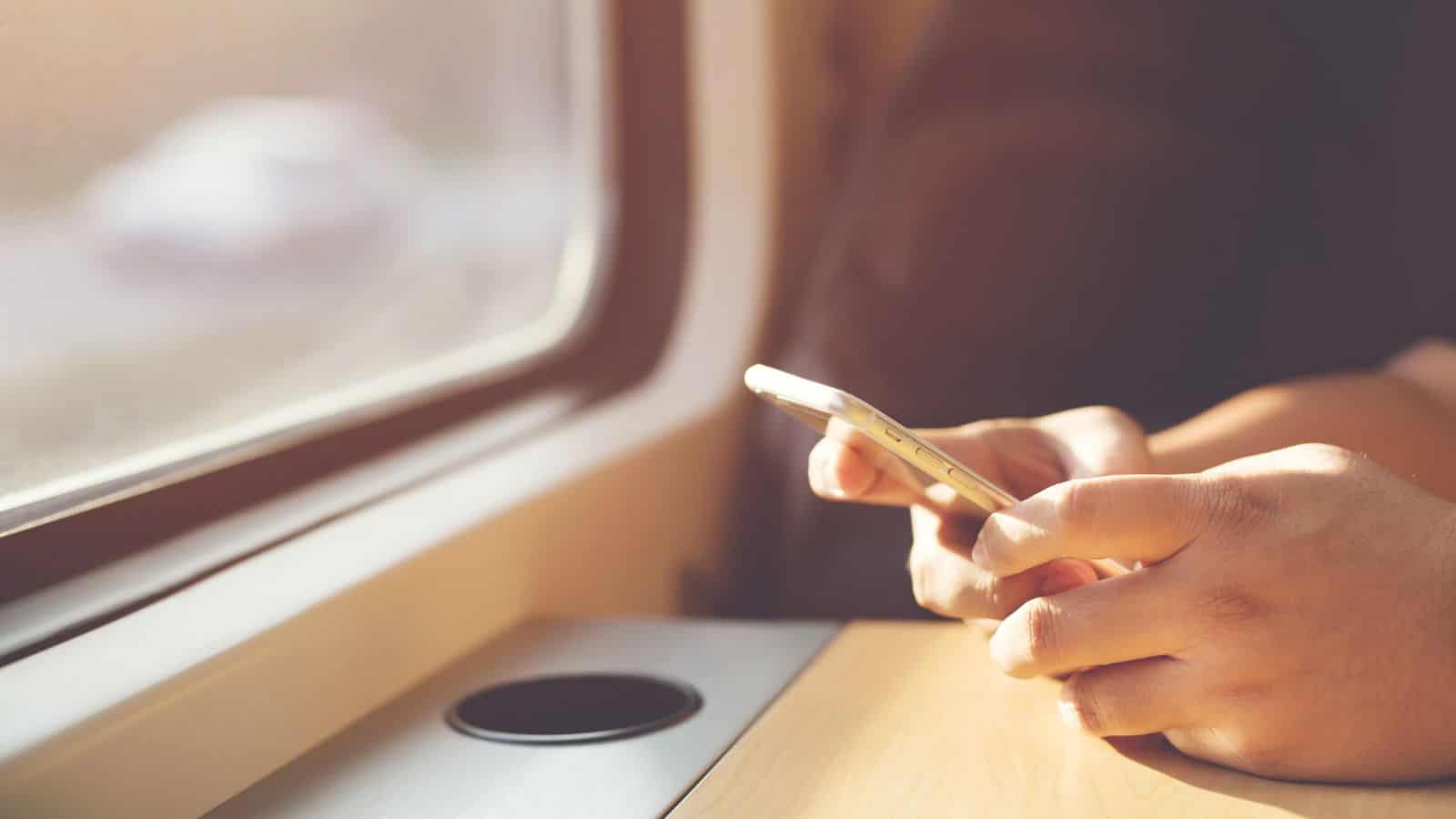 A person holds a smartphone with both hands while sitting next to a window on a train ride. Sunlight enters through the window, illuminating the scene.