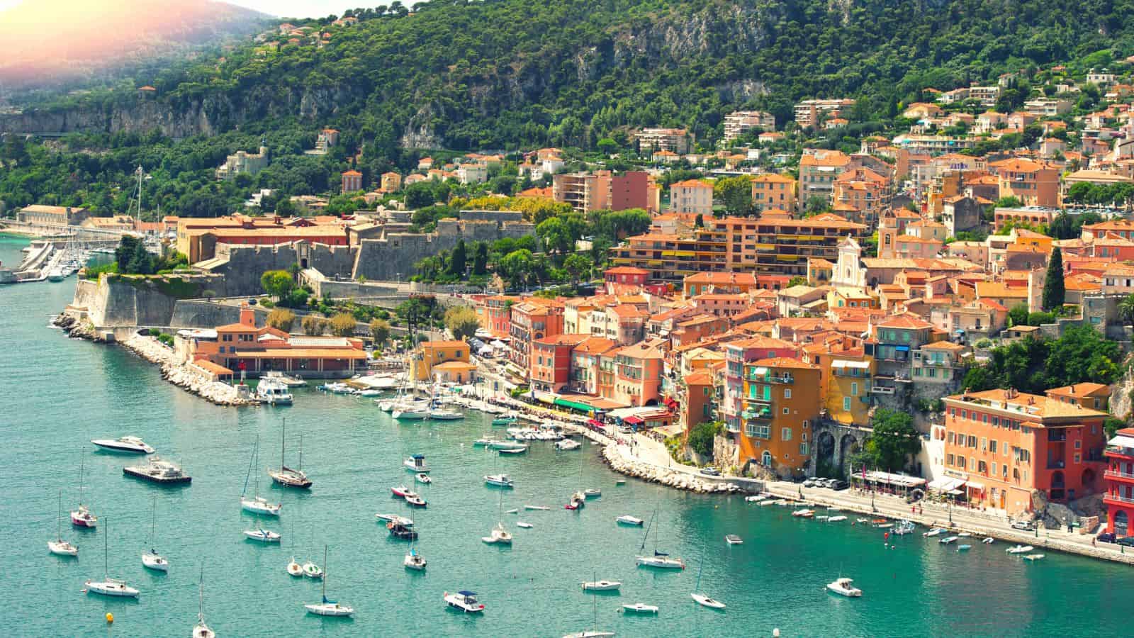 A coastal town with colorful buildings, boats in the harbor, and green hills in the background.