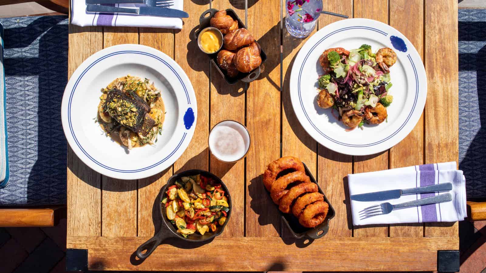 A wooden table set with two plates of food, a skillet of roasted vegetables, onion rings, a basket of bread rolls with dipping sauce, a glass of beer, and utensils on folded napkins offers travelers reasons to savor all-inclusive vacations.