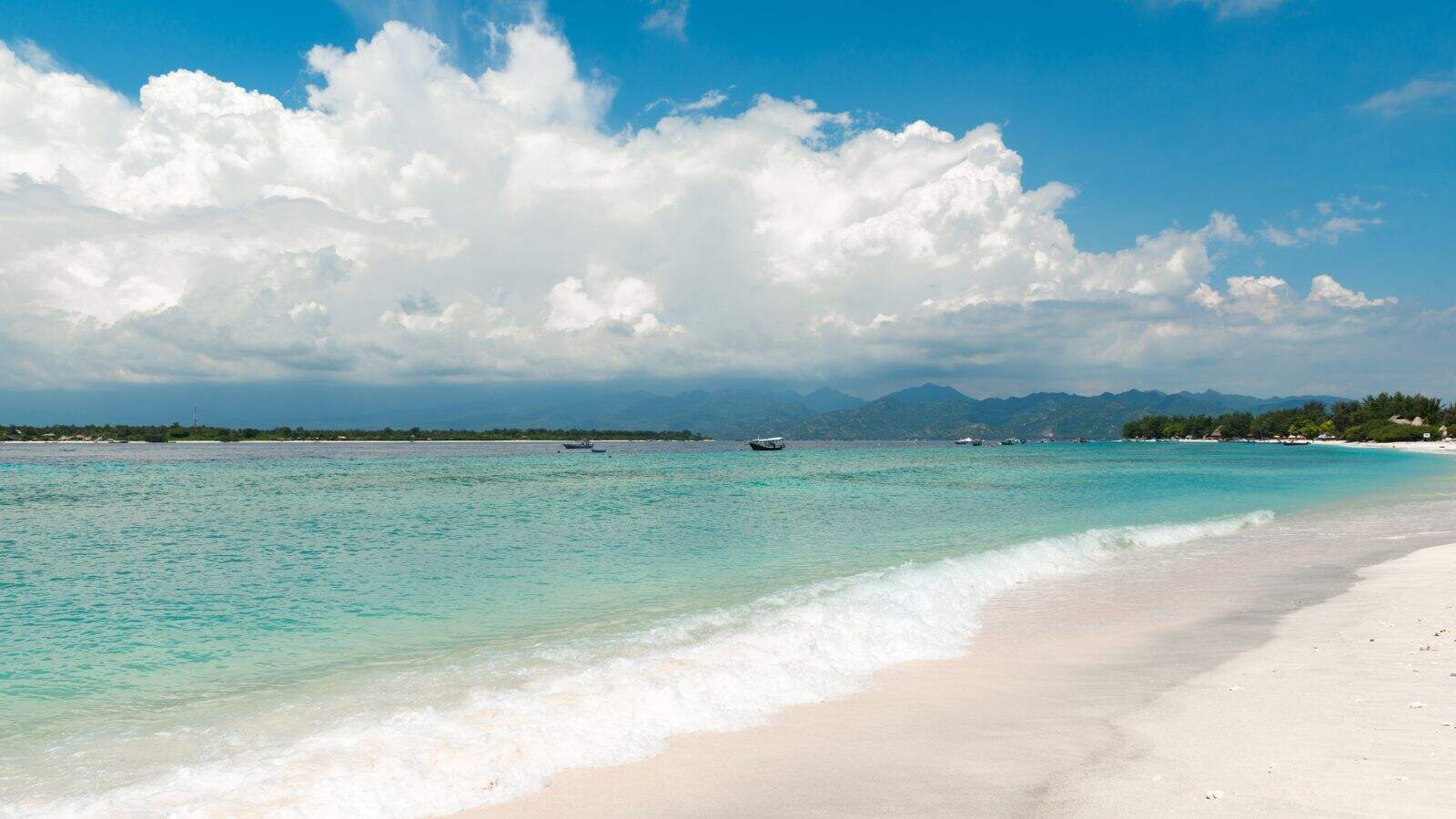 A white sandy beach with clear turquoise water under a partly cloudy sky. There are a few small boats on the water, and a distant shoreline with trees and mountains in the background.