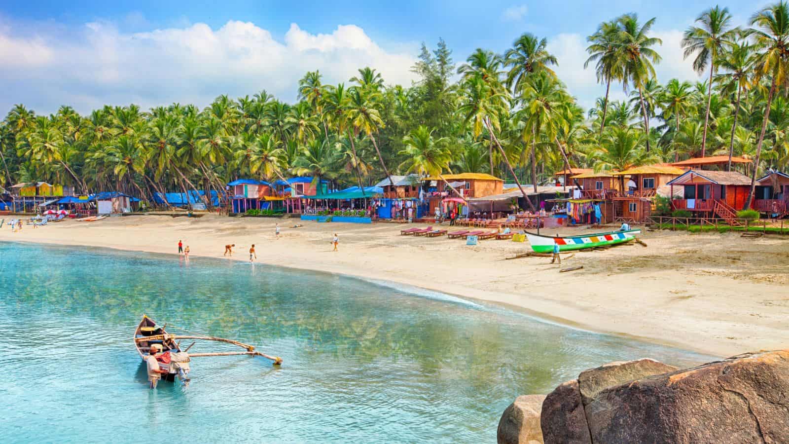 A sandy beach with colorful huts and palm trees along the shore, small boats near the water, and people walking and playing on the beach under a blue sky with scattered clouds.