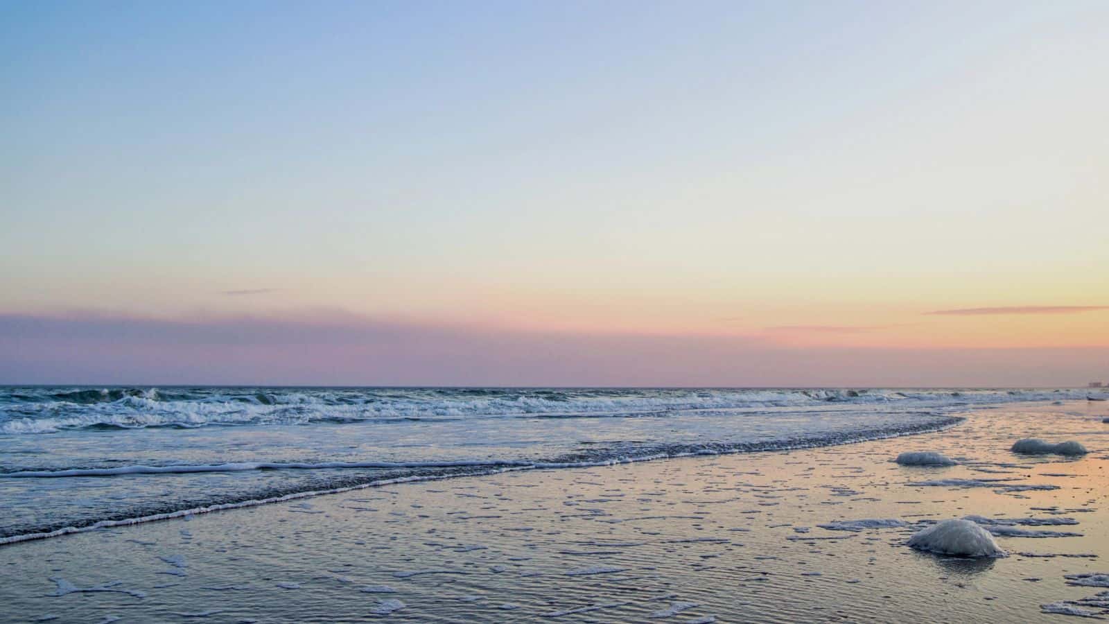 A calm beach scene at sunset with gentle waves rolling onto wet sand. The sky is gradient with soft pastel colors, and a few patches of foam are visible along the shoreline.