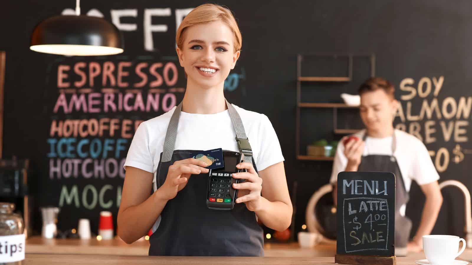 Smiling barista in apron processes card payment at coffee shop counter; menu board in background.