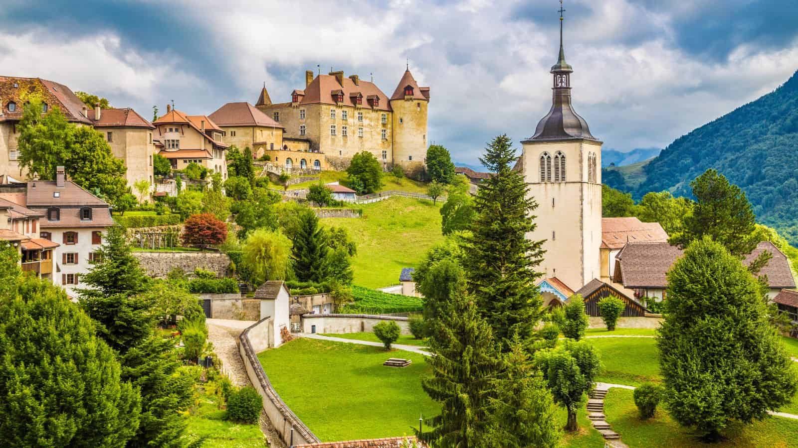 Medieval village with a castle and church tower surrounded by green hills and trees under a cloudy sky.