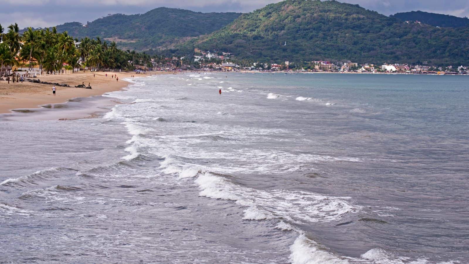 Waves roll onto a sandy beach lined with palm trees. A few people walk along the shoreline. Hills covered in greenery rise in the background, with buildings visible at the base near the water.