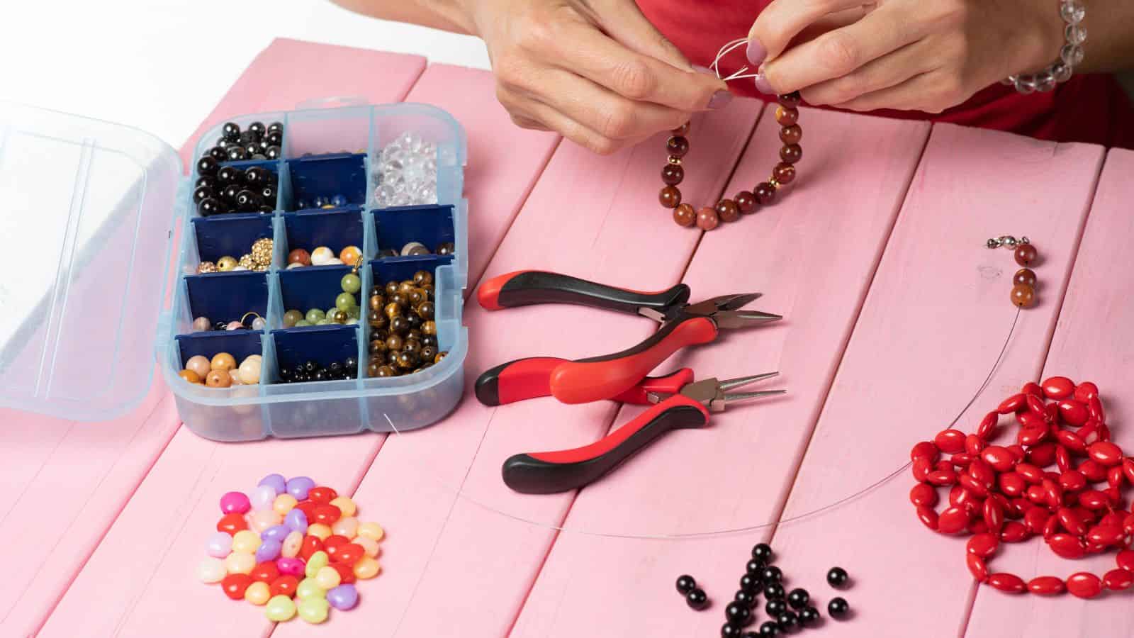 Hands making a beaded bracelet on a pink table with jewelry tools, beads, and a bead organizer.