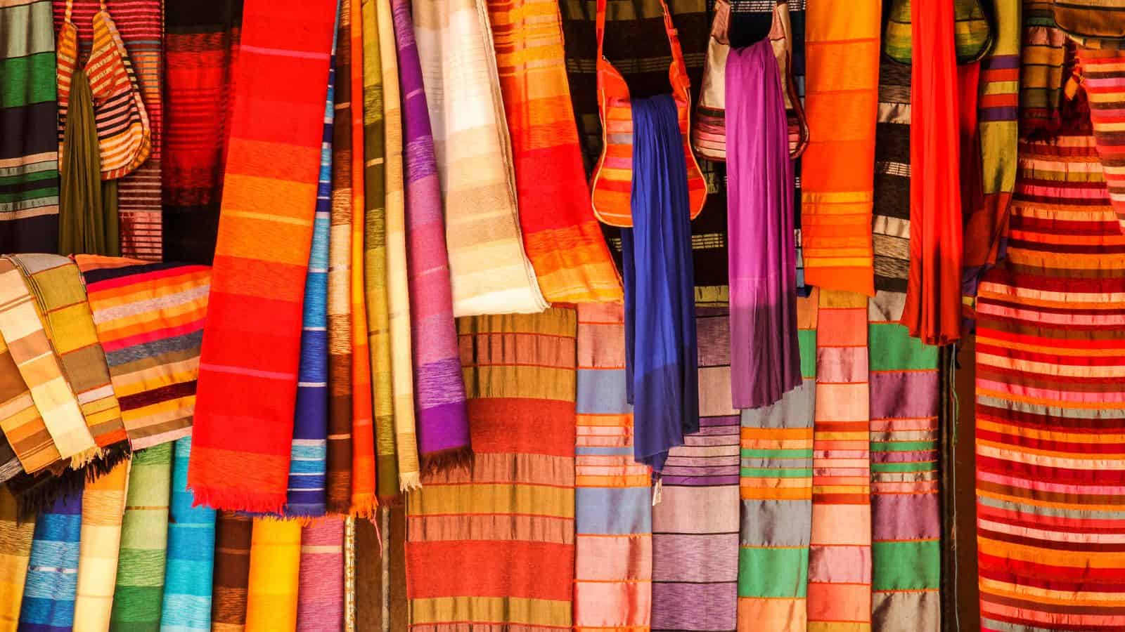 Colorful woven textiles and scarves with vibrant stripes and patterns hanging on display in a market.