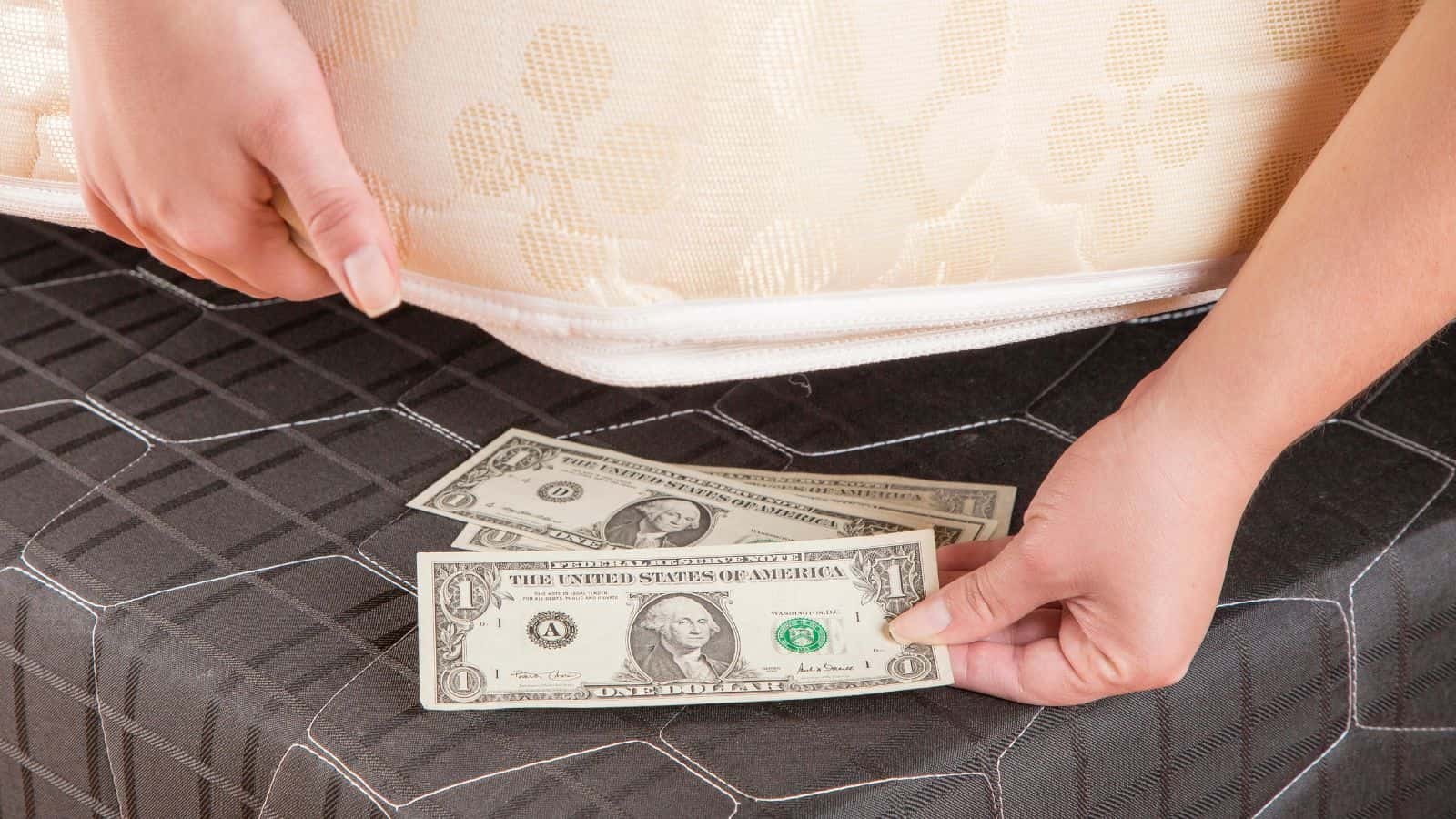Hands hiding dollar bills under a mattress with a patterned bedsheet.