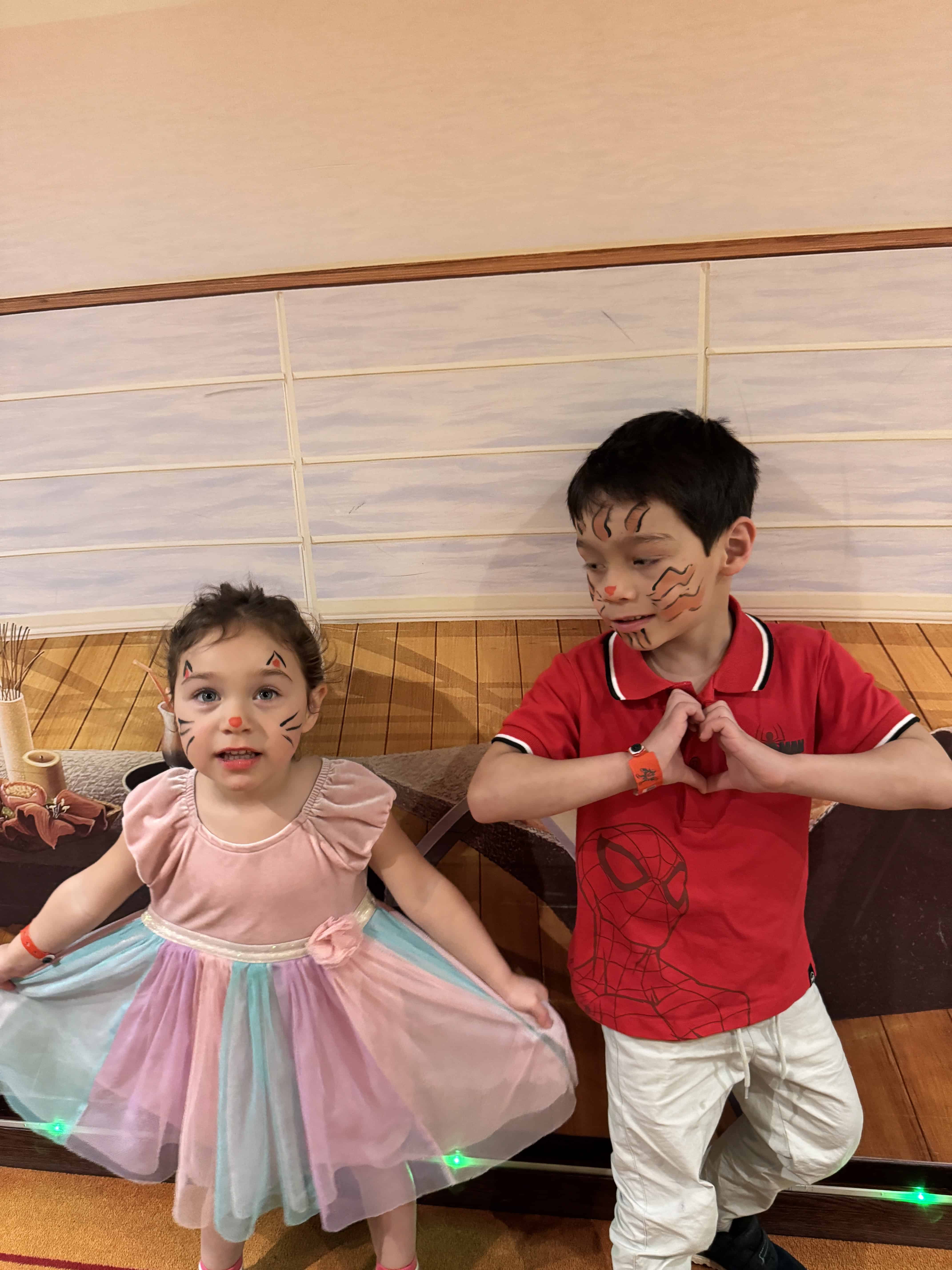 Two children with painted faces pose, one in a pastel dress and the other in a red shirt, against a wooden backdrop, capturing memories from their MSC Cruise Fantasia adventure.
