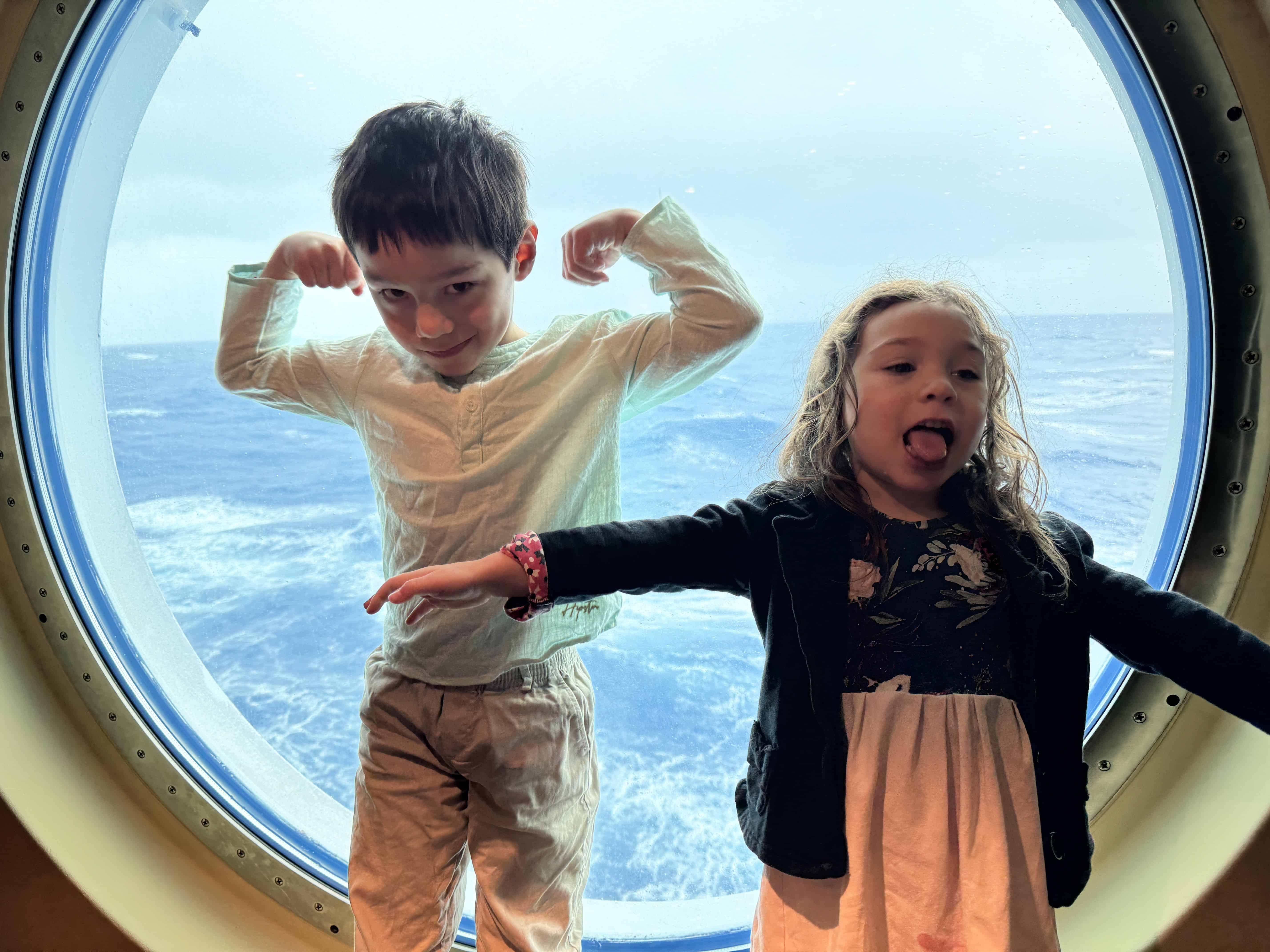 Two kids pose playfully in front of a round ship window aboard the MSC Cruise Fantasia, with the ocean in the background.