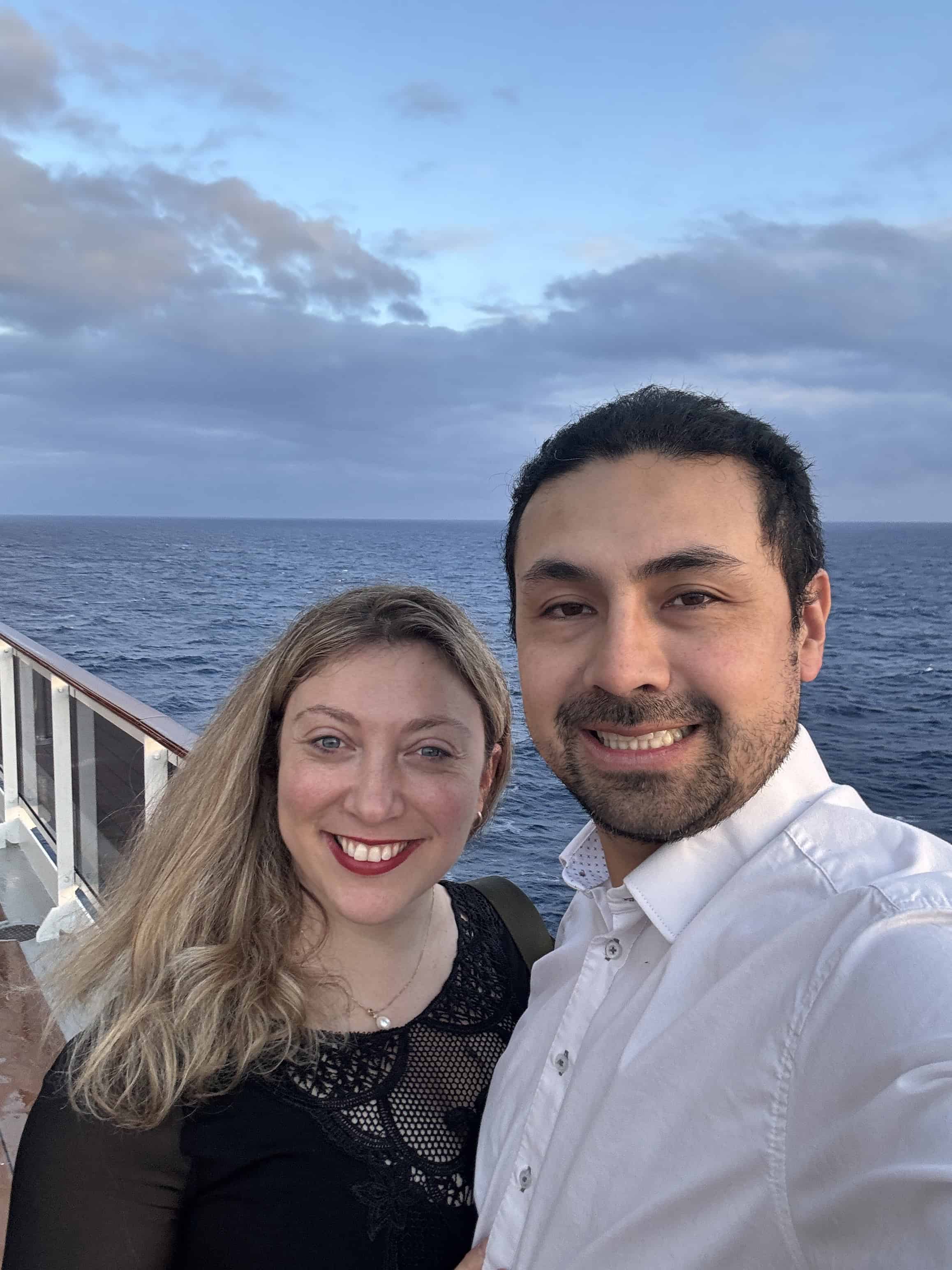 A smiling couple takes a selfie on the deck of MSC Cruise Fantasia, with the ocean and cloudy sky in the background.